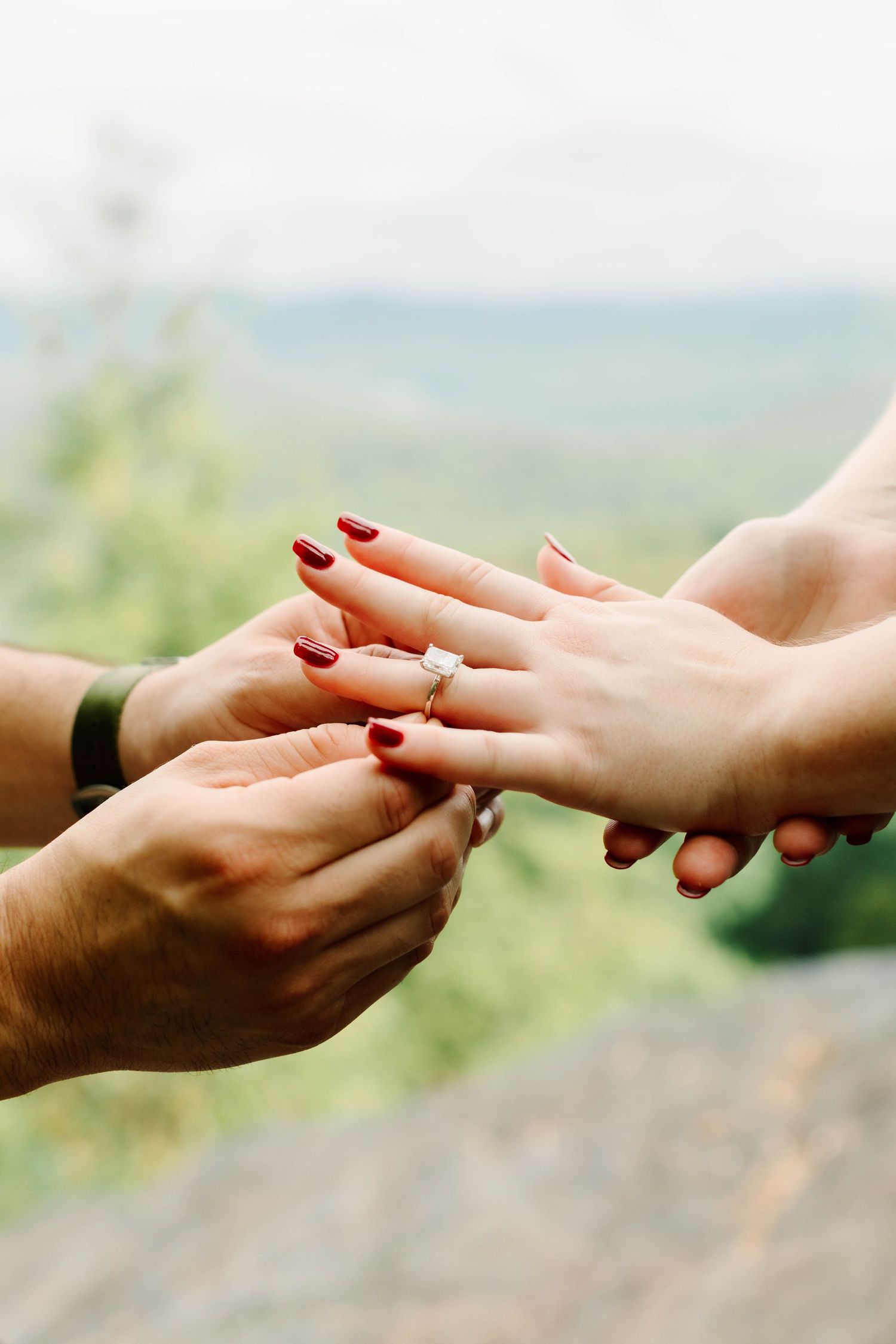 Close-up of man placing diamond engagement ring on womans hand with red nails against mountain backdrop.