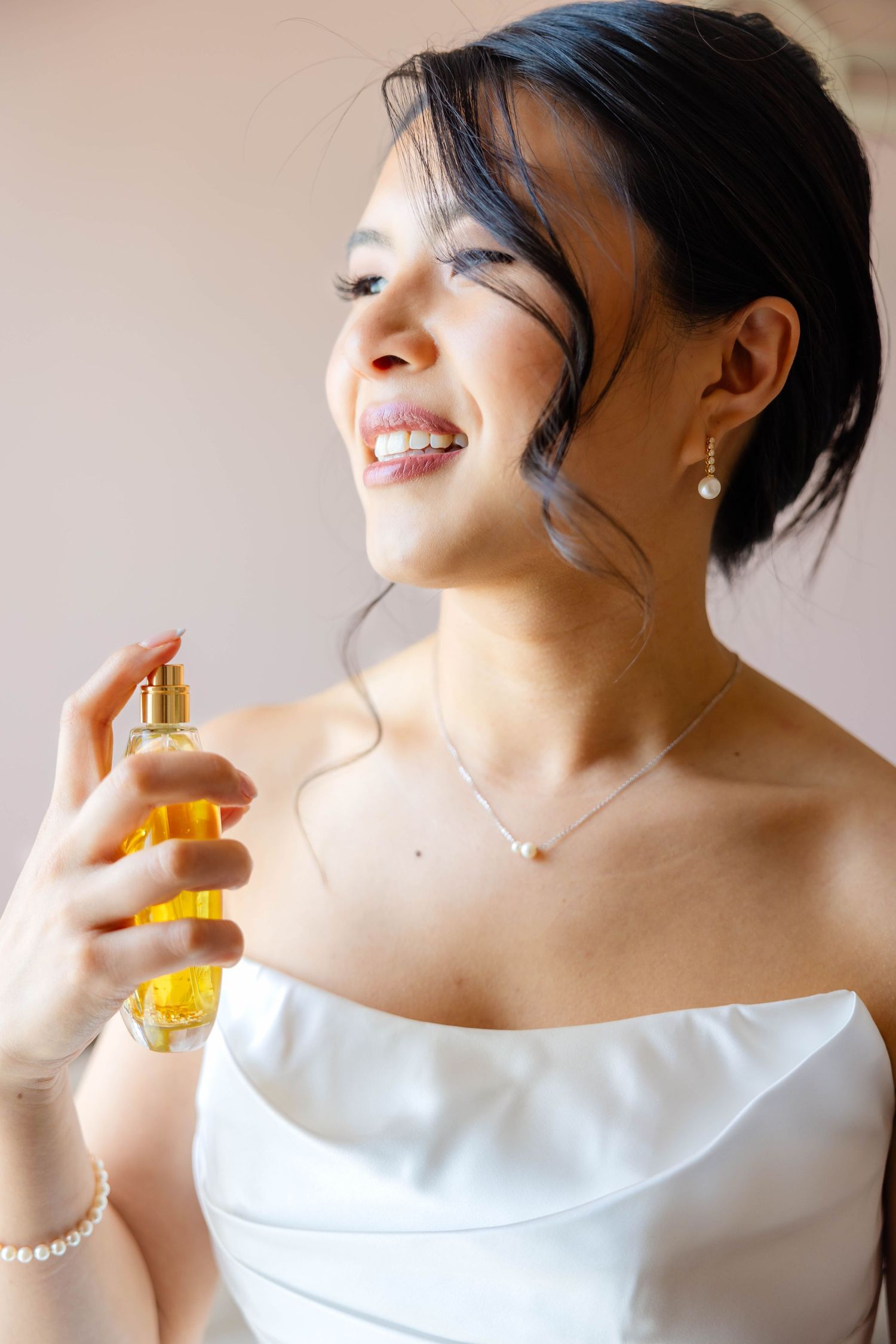 Smiling bride in white strapless dress applying perfume with pearl earrings and necklace.