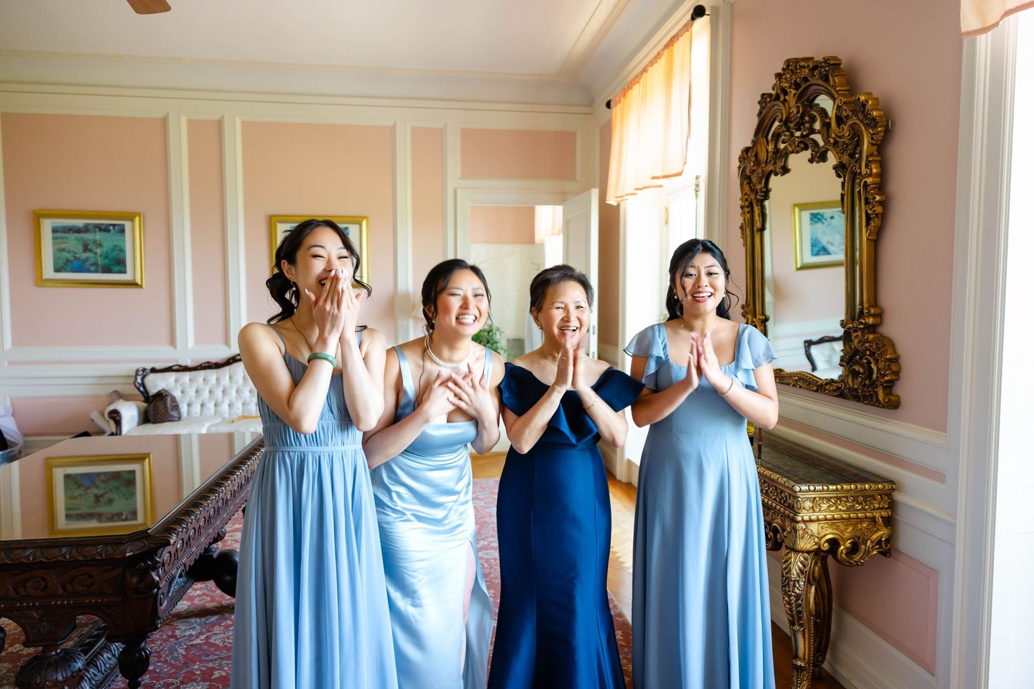 Four bridesmaids in blue gowns react with joy and excitement while getting ready in an elegant pink room.