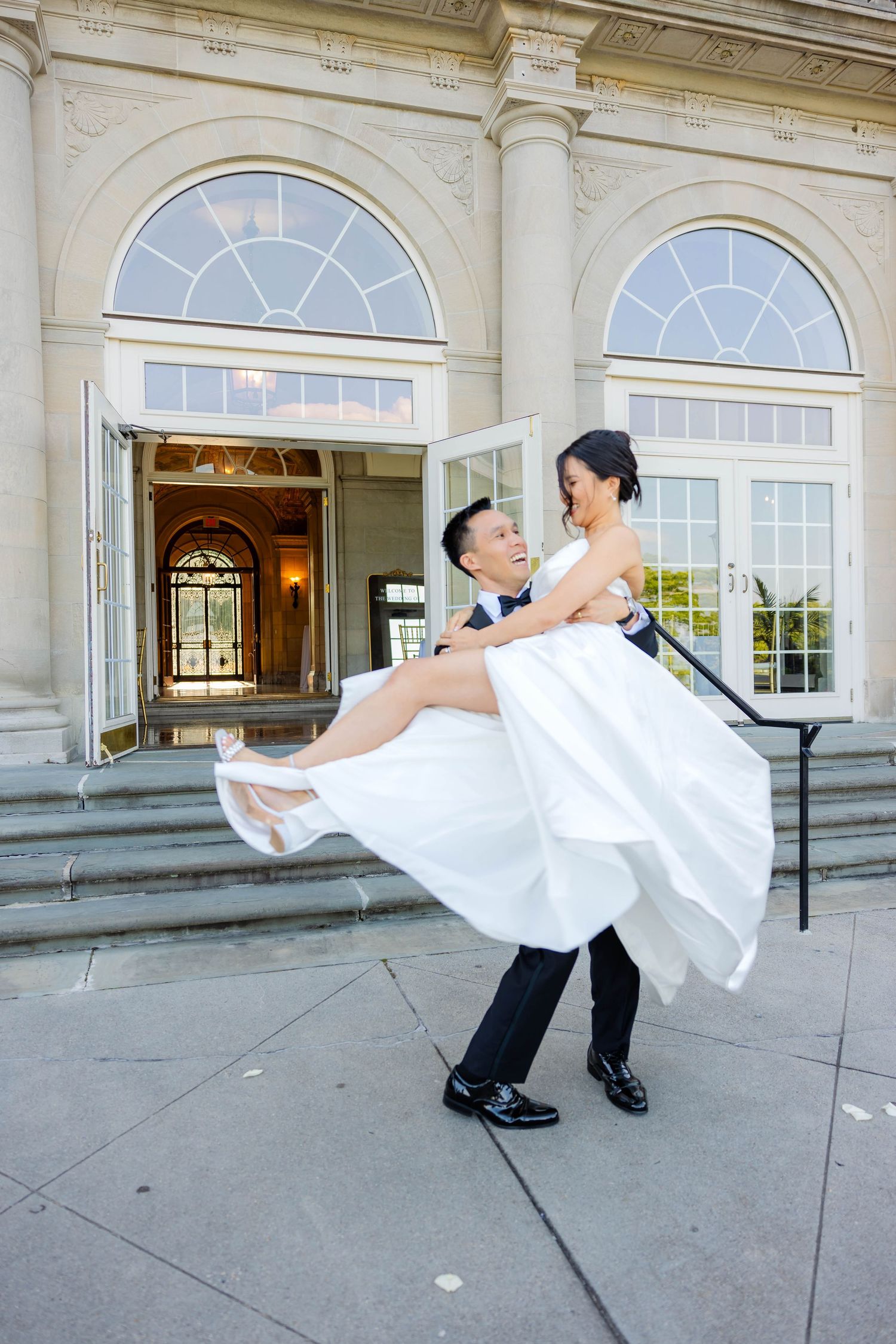 Joyful groom in black tuxedo lifting smiling bride in white wedding dress outside elegant venue entrance.