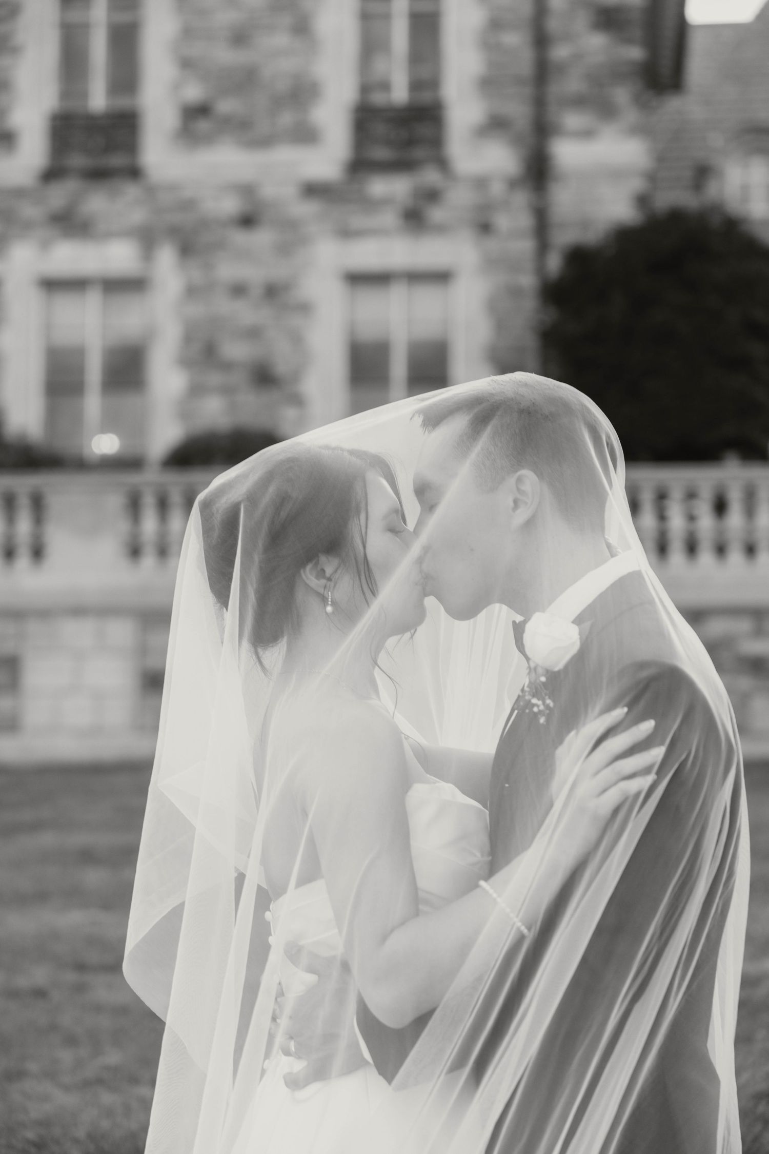 Black and white photo of bride and groom kissing under wedding veil outside historic stone building.