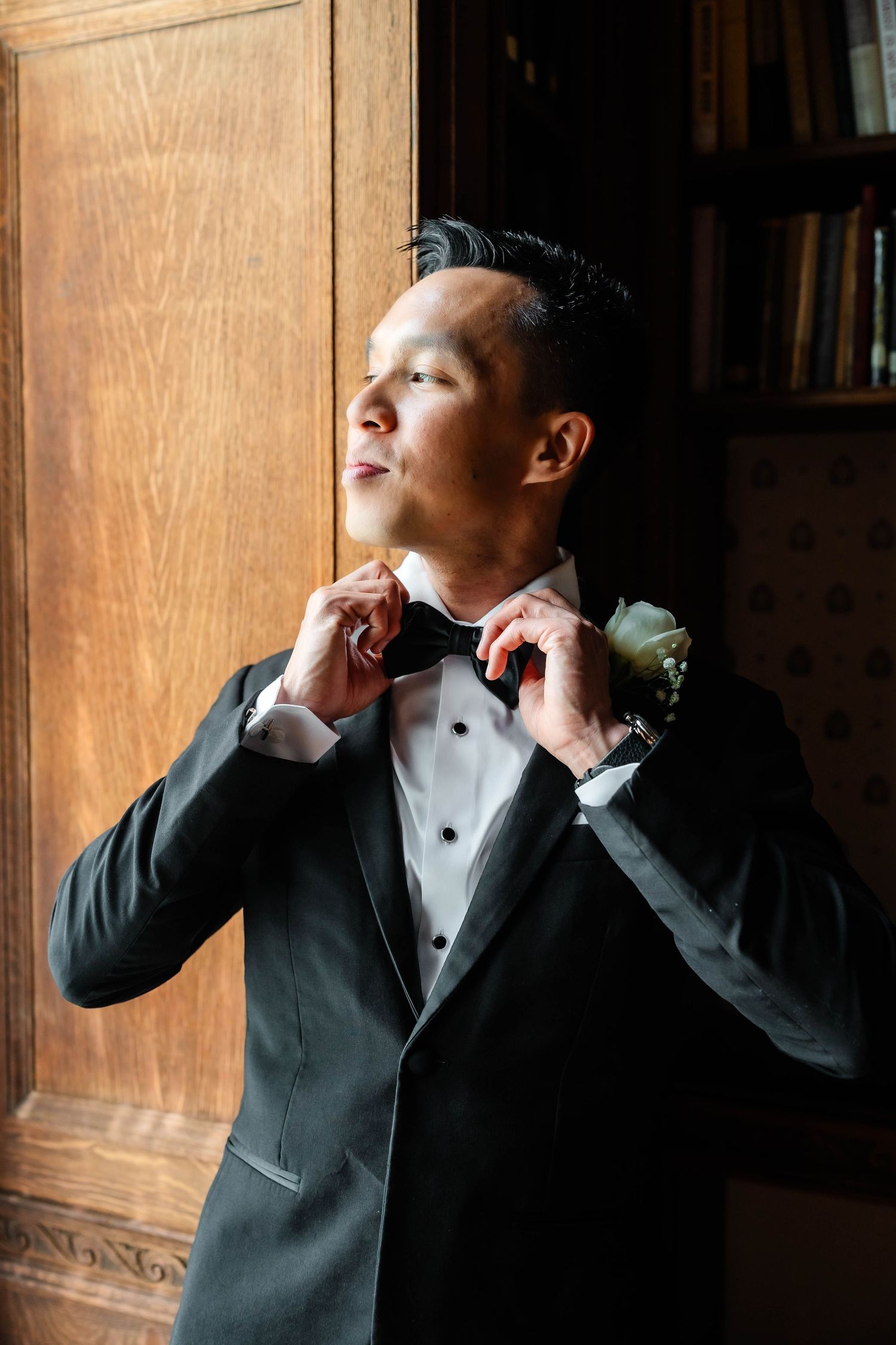 Dapper groom in black tuxedo adjusts bow tie with white rose boutonniere beside wooden bookcase.