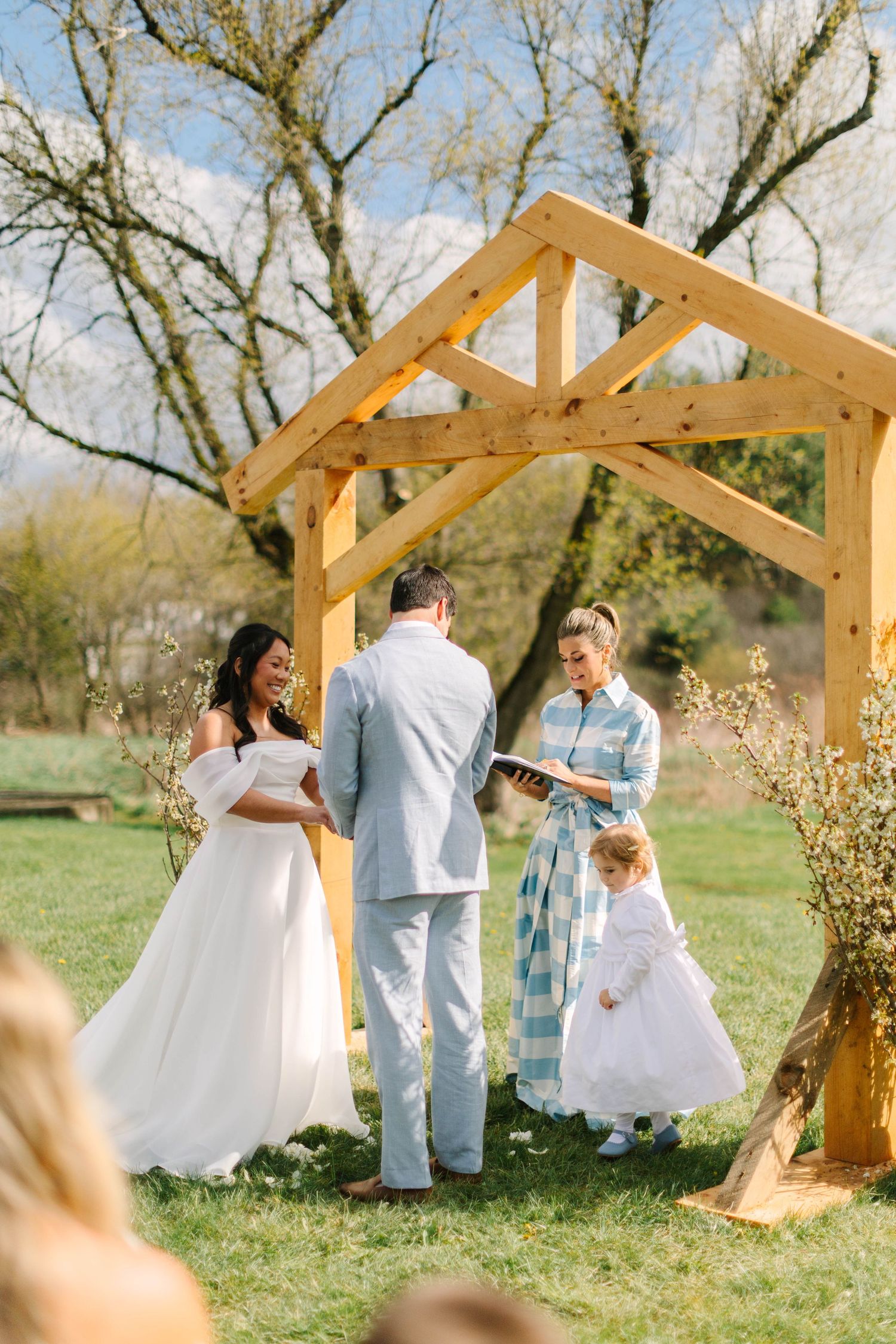 Couple exchanges vows under wooden arch with officiant and flower girl at outdoor spring wedding.