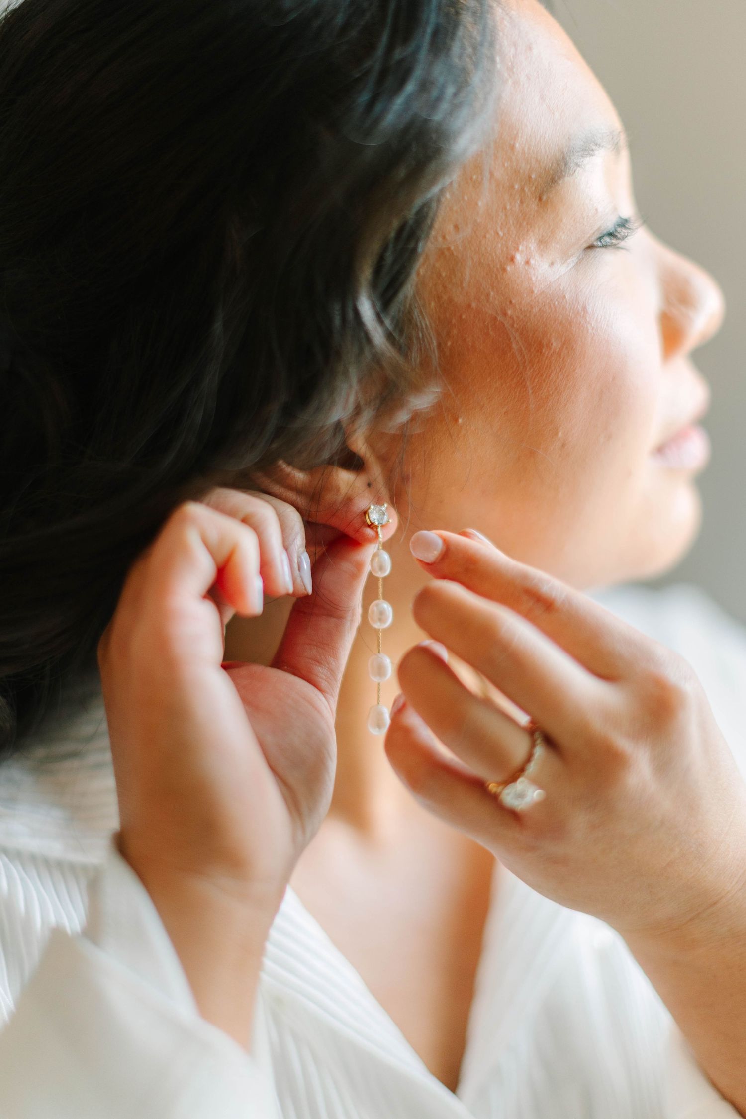 Bride with dark hair puts on delicate crystal drop earrings while wearing engagement ring.