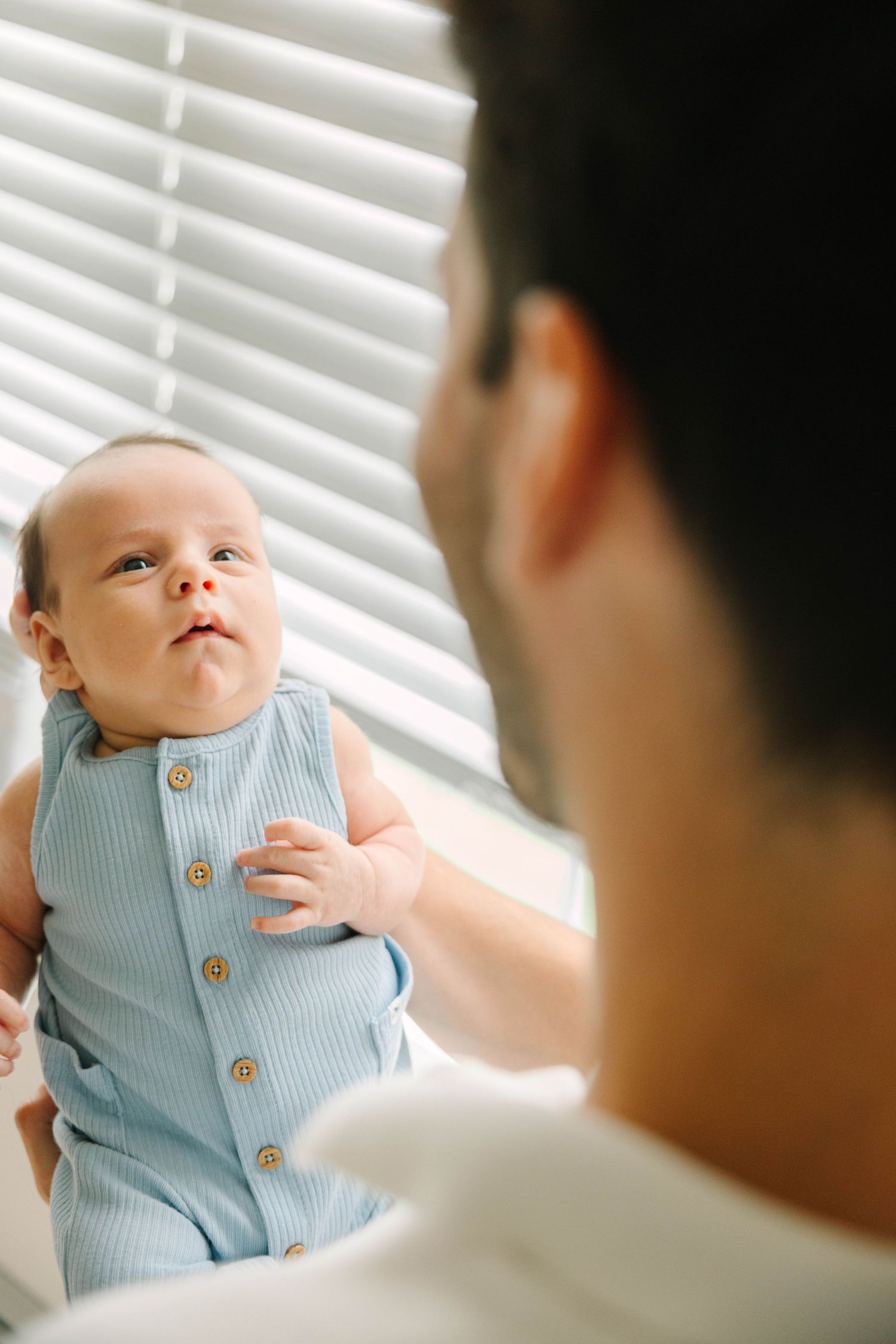 A father holds his newborn baby in a blue ribbed onesie near a window with white blinds.