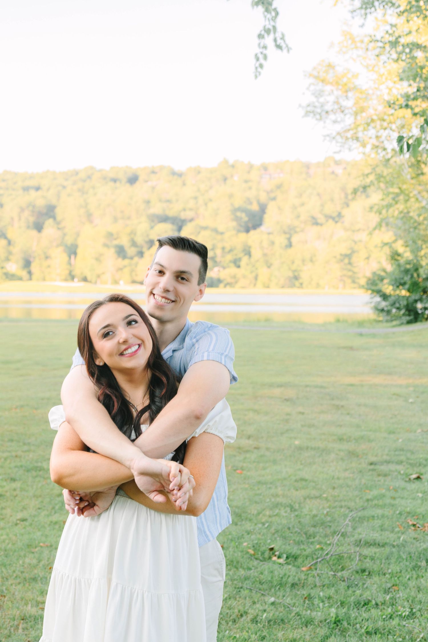 Man hugging woman from behind in a sunny green park near a lake.