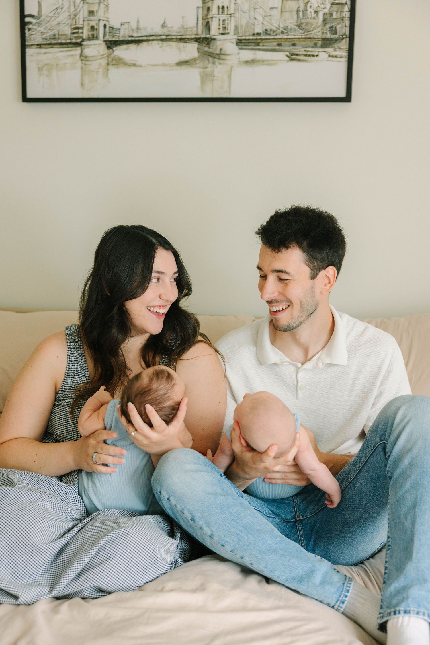 Happy young parents sitting on a bed smiling at each other while holding their newborn baby.