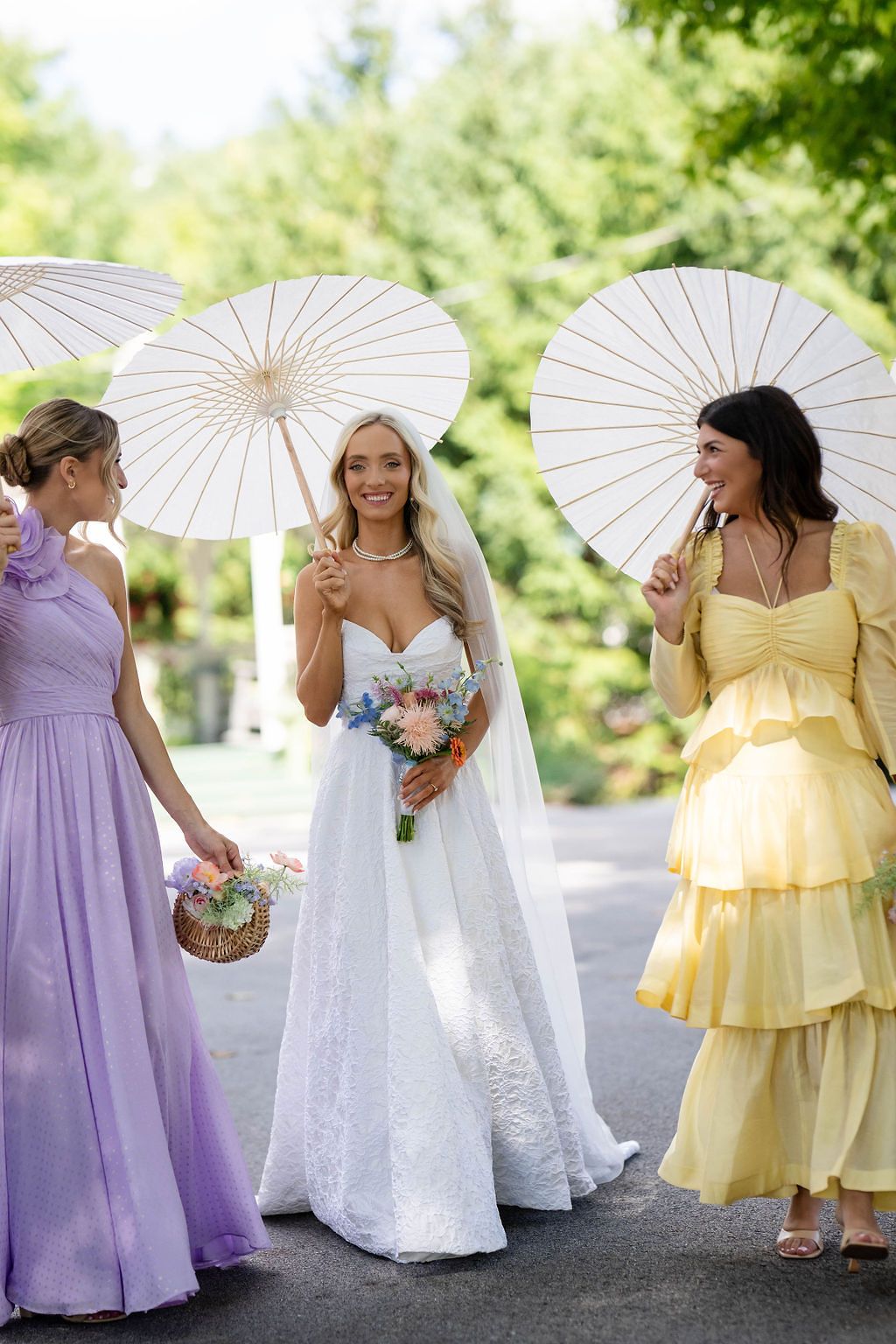 Bride in white gown with two bridesmaids in purple and yellow dresses holding white parasols outdoors.