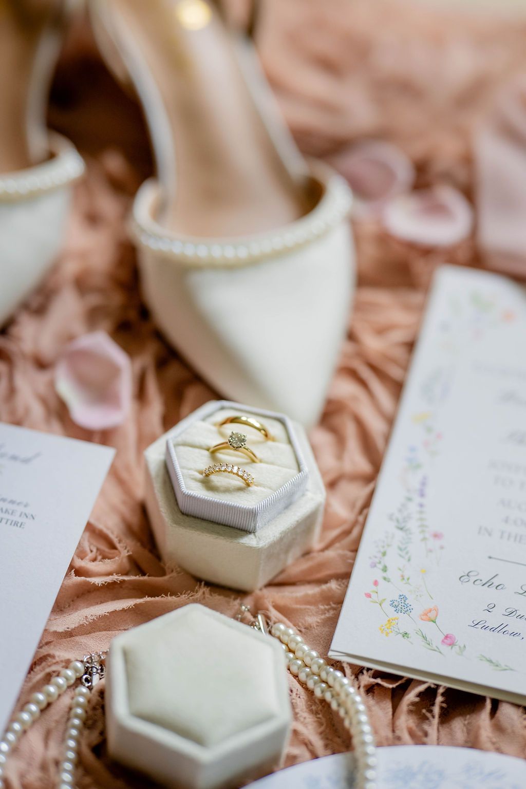 Wedding flat lay with gold rings in velvet box, pearl necklace, floral invitation, and ivory heels on blush fabric.