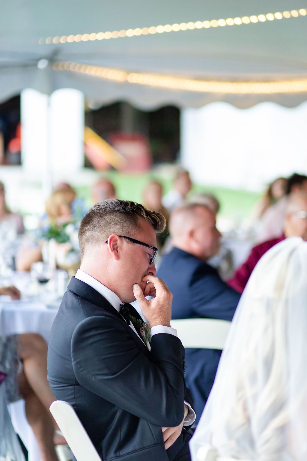 Groom in dark suit listens emotionally to wedding speeches at a tented outdoor reception.