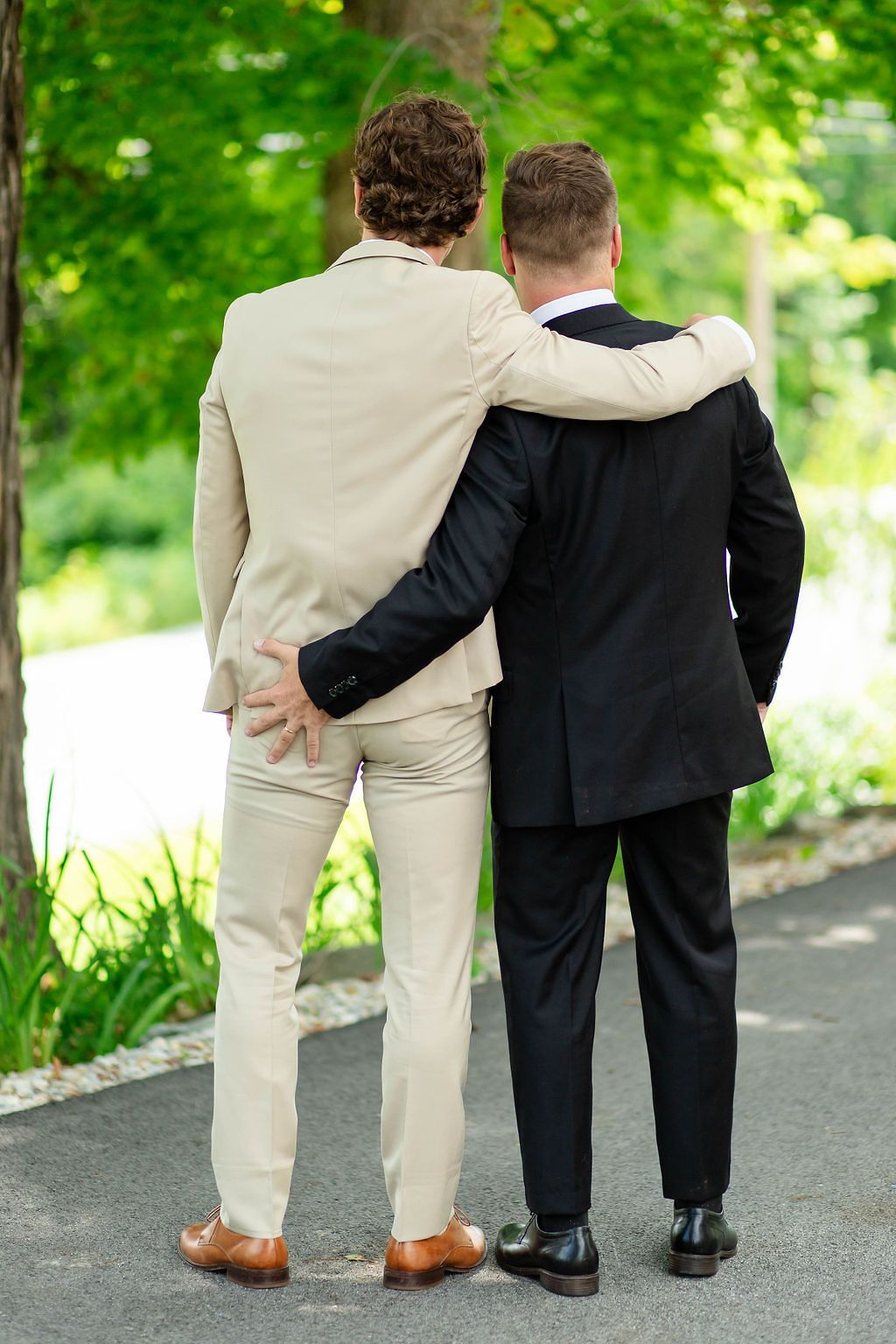 Two grooms in suits walk arm in arm down a tree-lined path at their outdoor wedding.