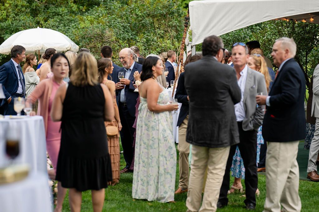 Wedding cocktail hour guests mingling outdoors under a white tent surrounded by lush green trees.