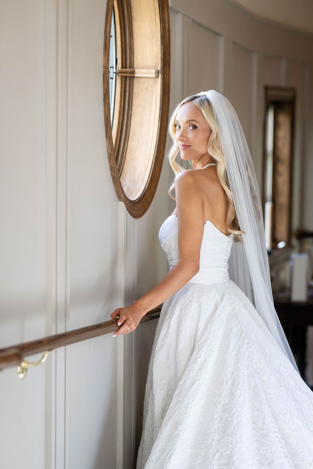 Elegant bride in strapless lace ball gown and veil poses on staircase beside round wooden mirror.
