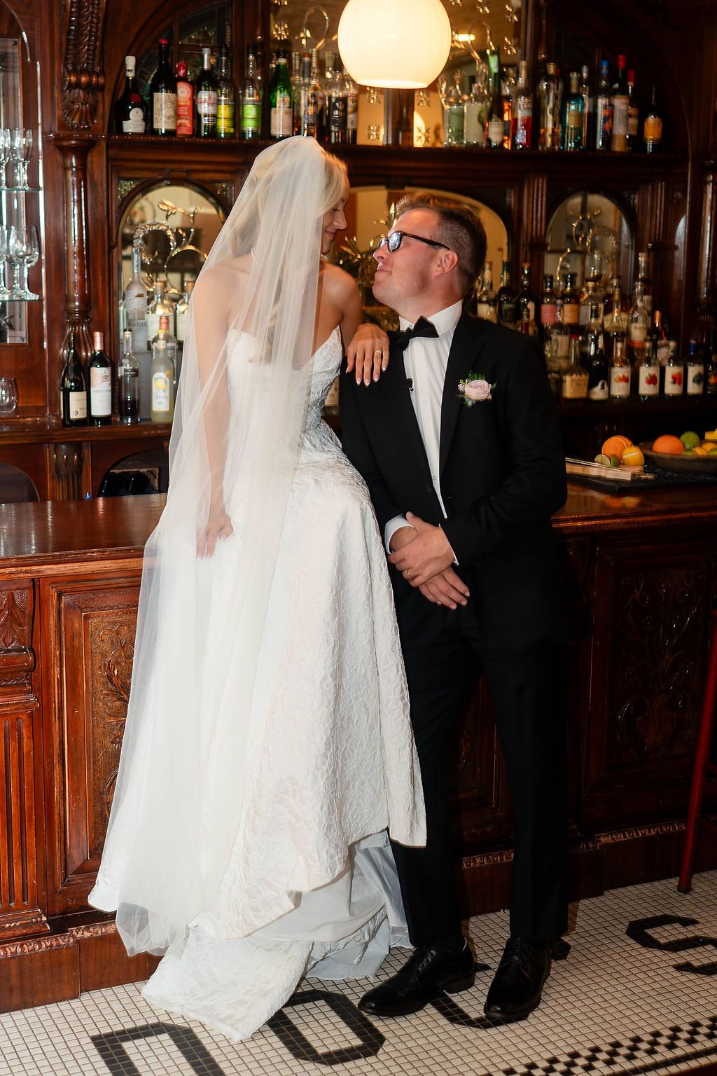 Bride in white lace gown and veil sharing a romantic moment with groom at a vintage bar.