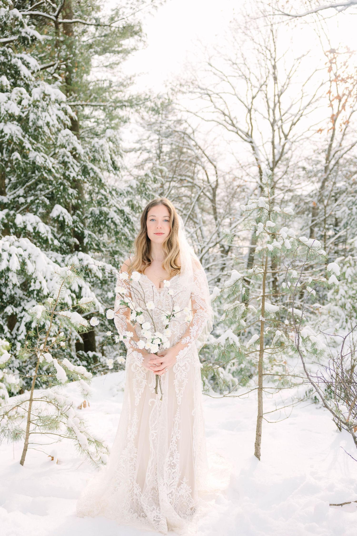 Bride in lace gown holding white bouquet stands in snowy winter forest with snow-covered pine trees.