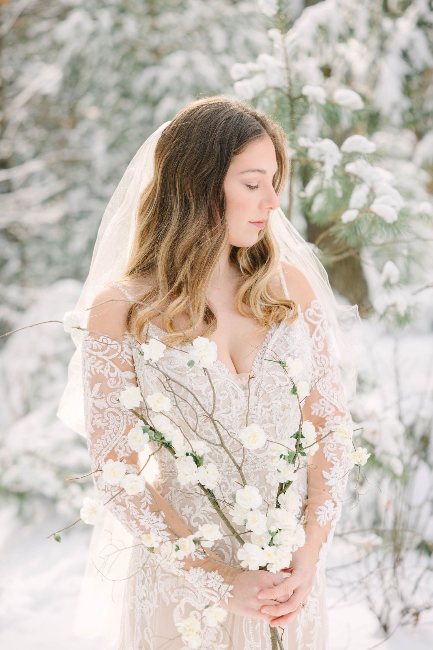 Bride in lace off-shoulder gown holding white cherry blossom branches in a snowy winter forest setting.
