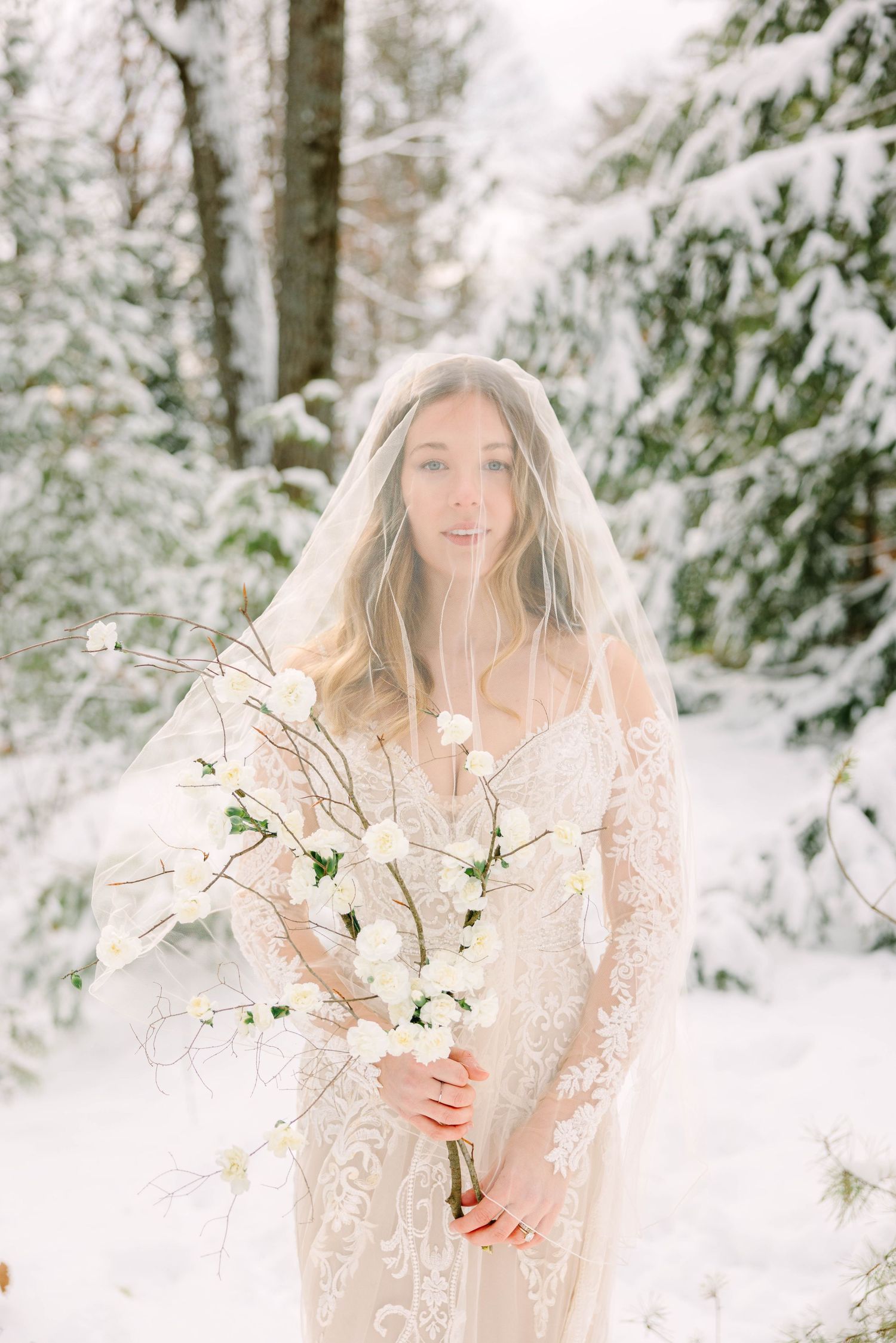 Bride in lace gown draped in veil holds white floral bouquet while standing among snow-covered evergreen trees.