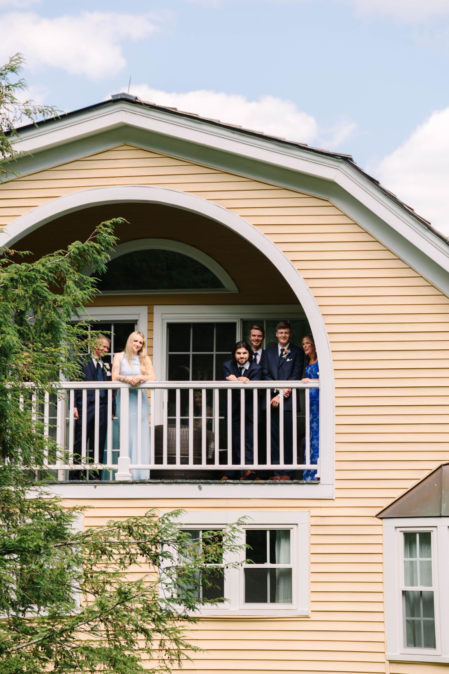Wedding party gathers on a balcony of a yellow barn-style venue under a blue sky.