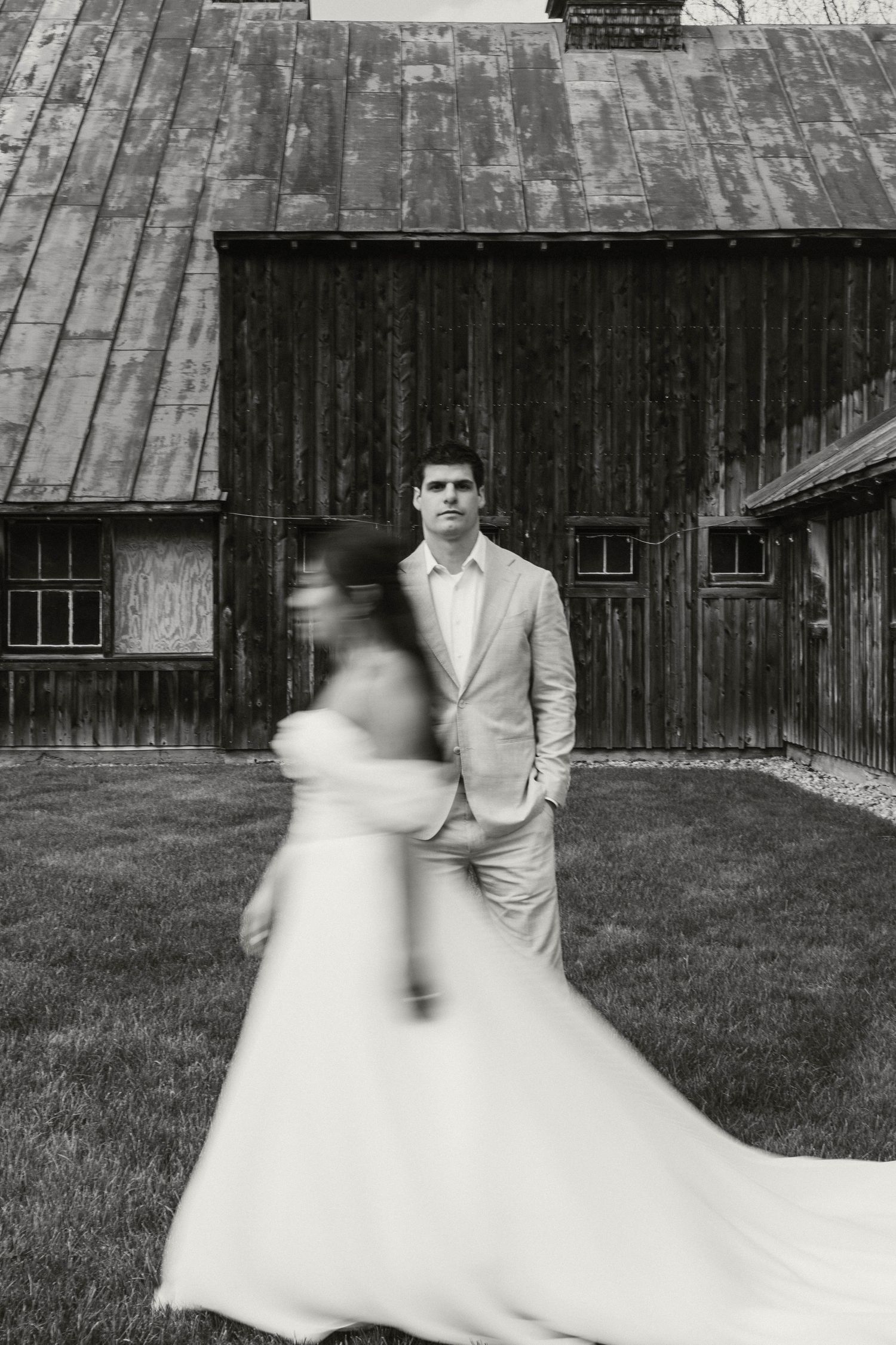 Black and white photo of groom standing before rustic barn as bride in flowing white gown walks toward him.