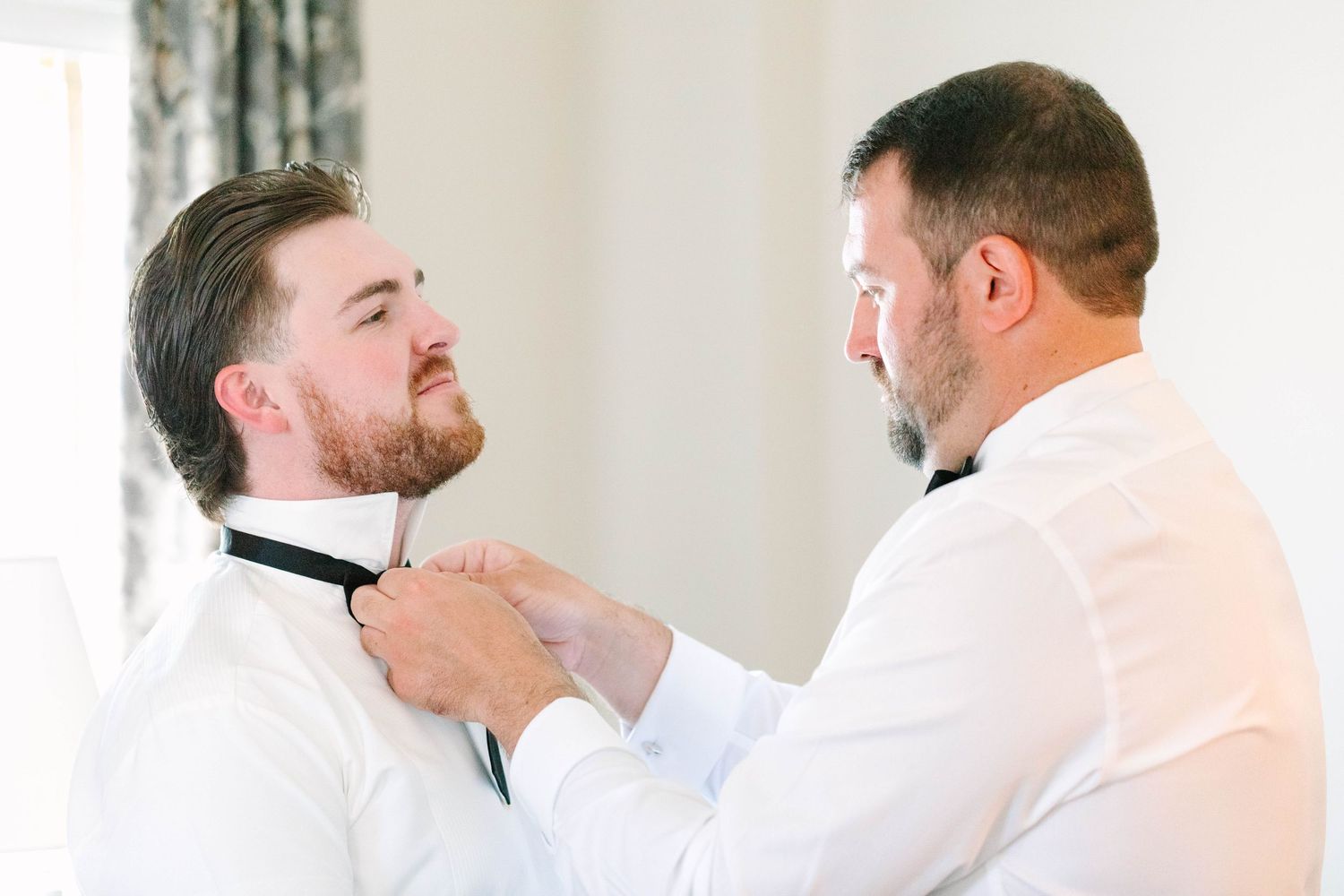 Man helping groom adjust his black bow tie while both wear white dress shirts before wedding.