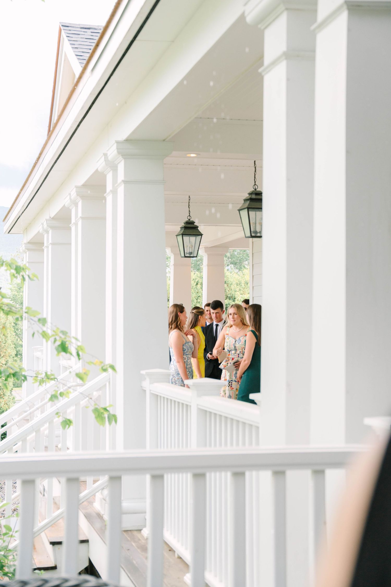 Wedding guests mingling on elegant white columned porch with hanging lanterns at a classic venue.