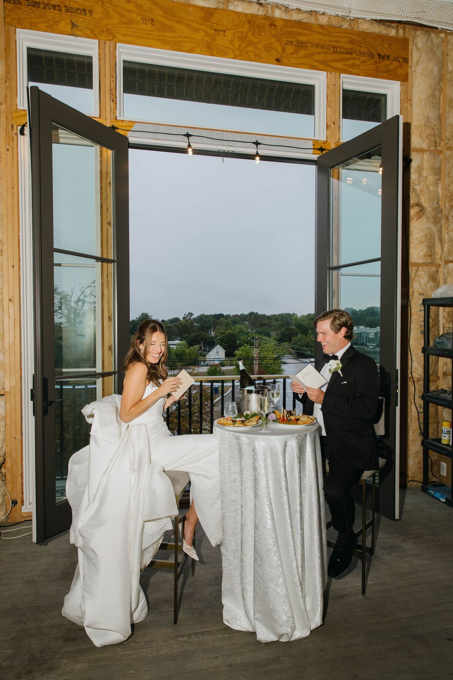 Bride and groom sit at a high-top table reading vows by open French doors with scenic outdoor views.
