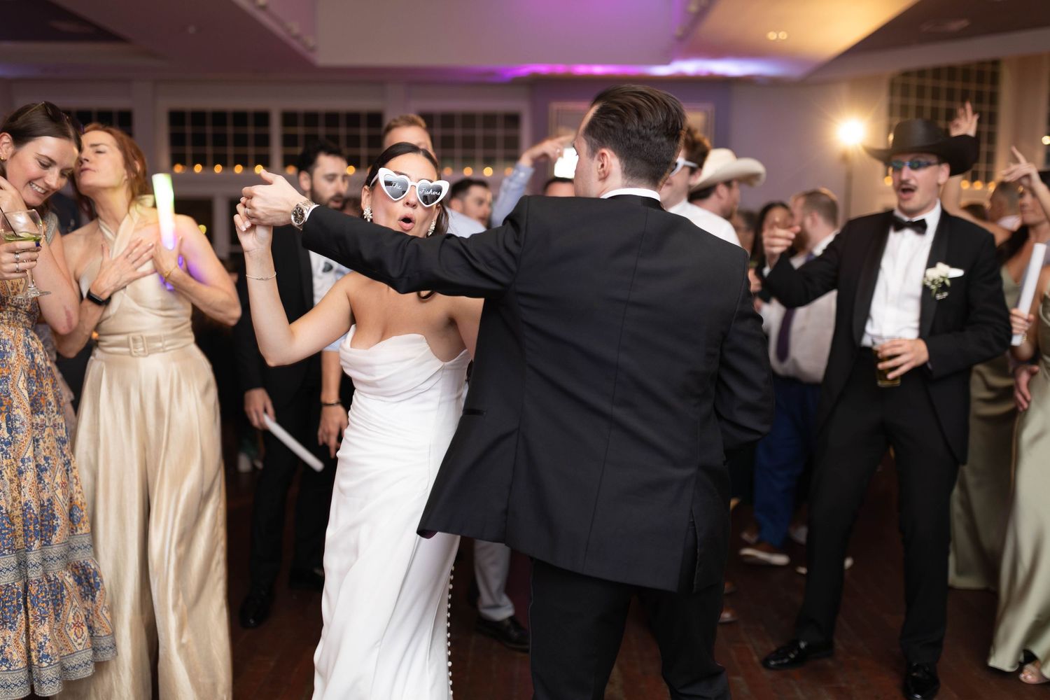 Bride and groom dance joyfully with guests wearing novelty sunglasses on crowded wedding reception dance floor.