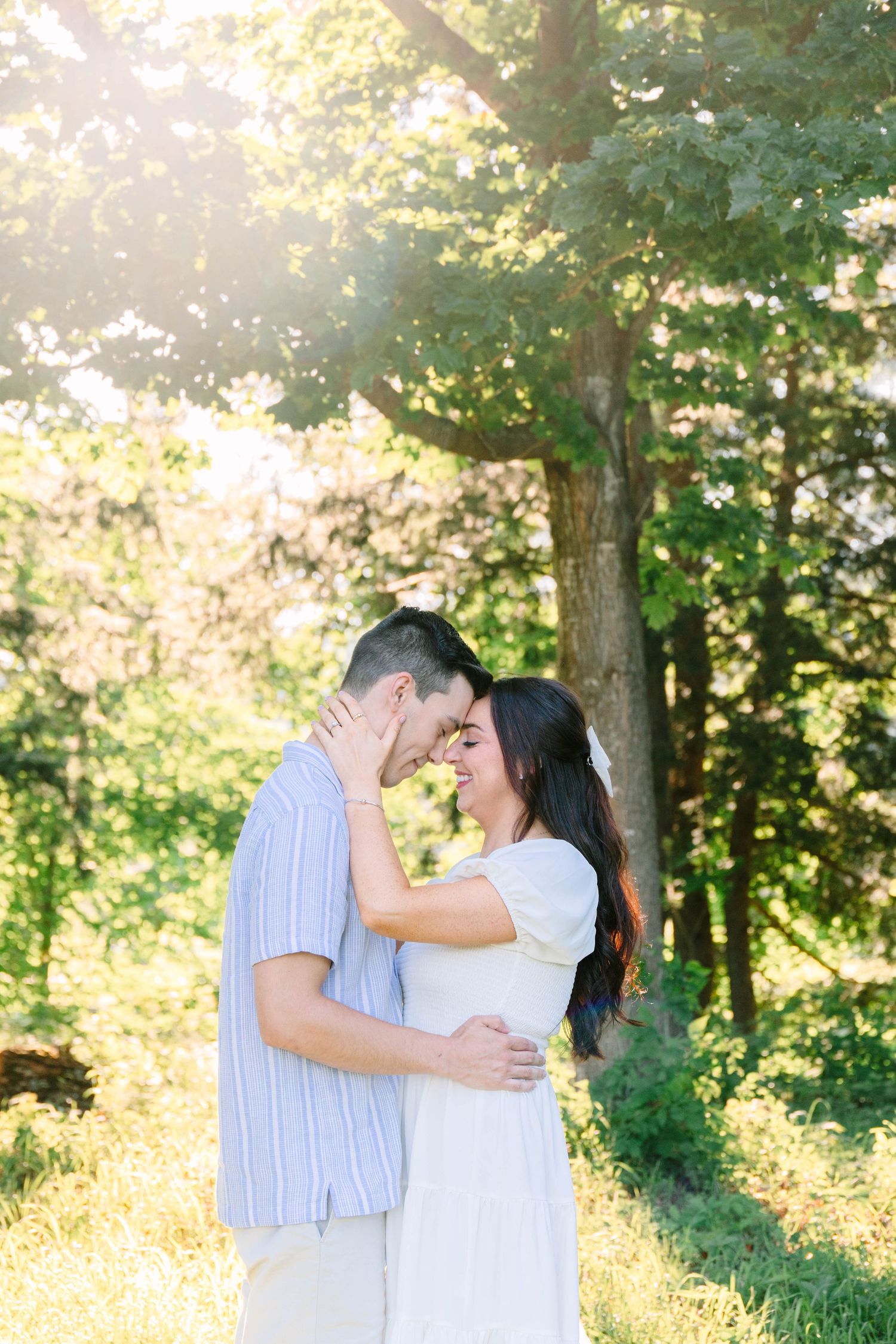 A couple shares a romantic forehead touch in a sunlit forest clearing, the woman in white dress and man in striped shirt embracing.