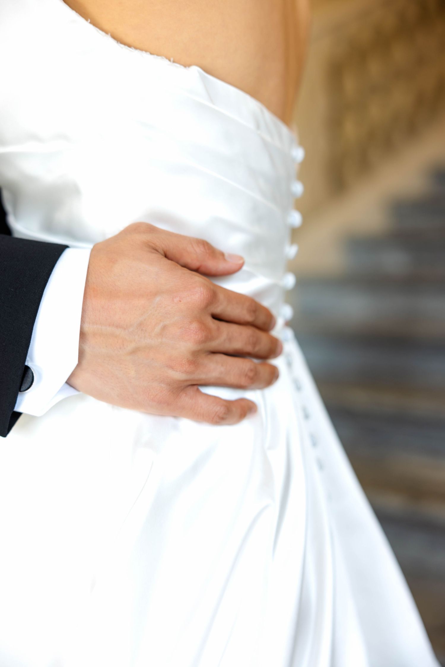 Close-up of groom's hand gently holding bride's waist in white satin strapless wedding dress.