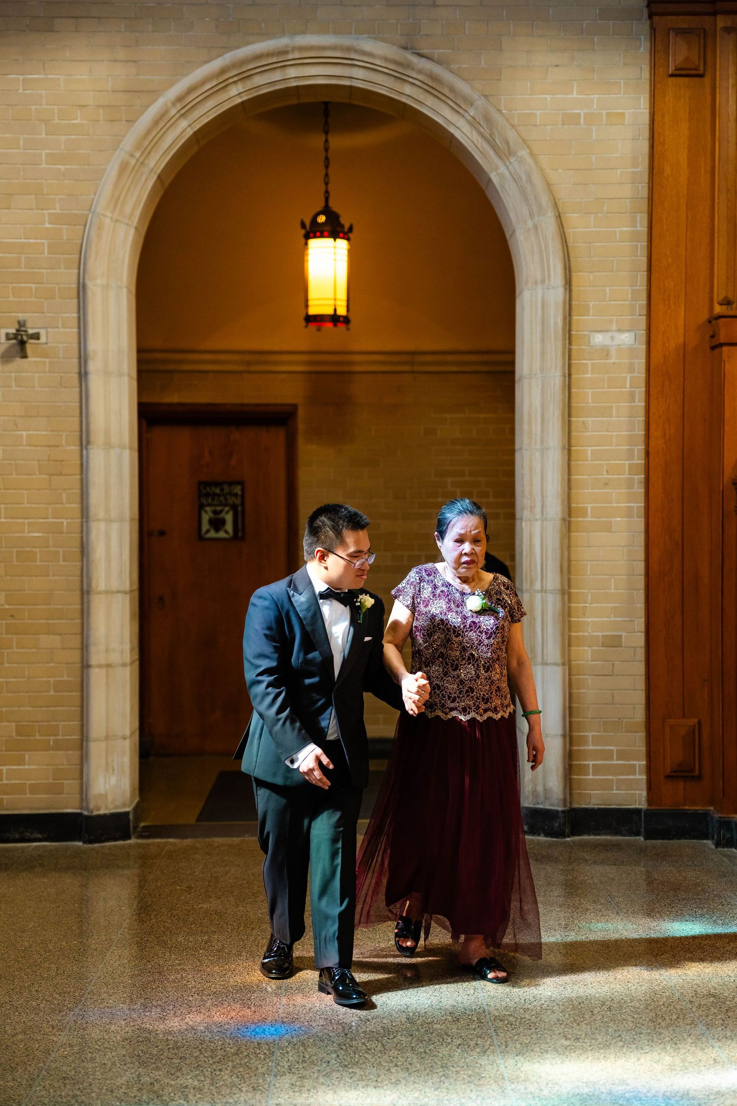 Groom in black tuxedo escorting woman in burgundy gown through arched church entrance with pendant light.