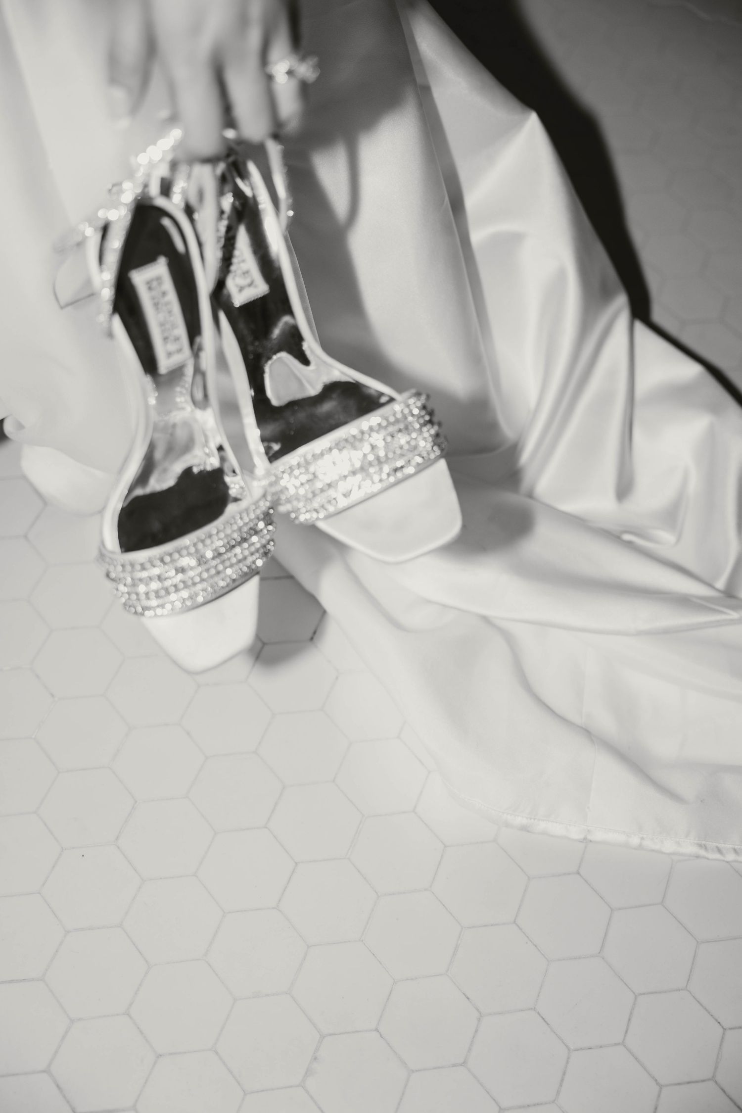 Black and white close-up of crystal-embellished white wedding heels on hexagonal tile floor beside bridal gown.