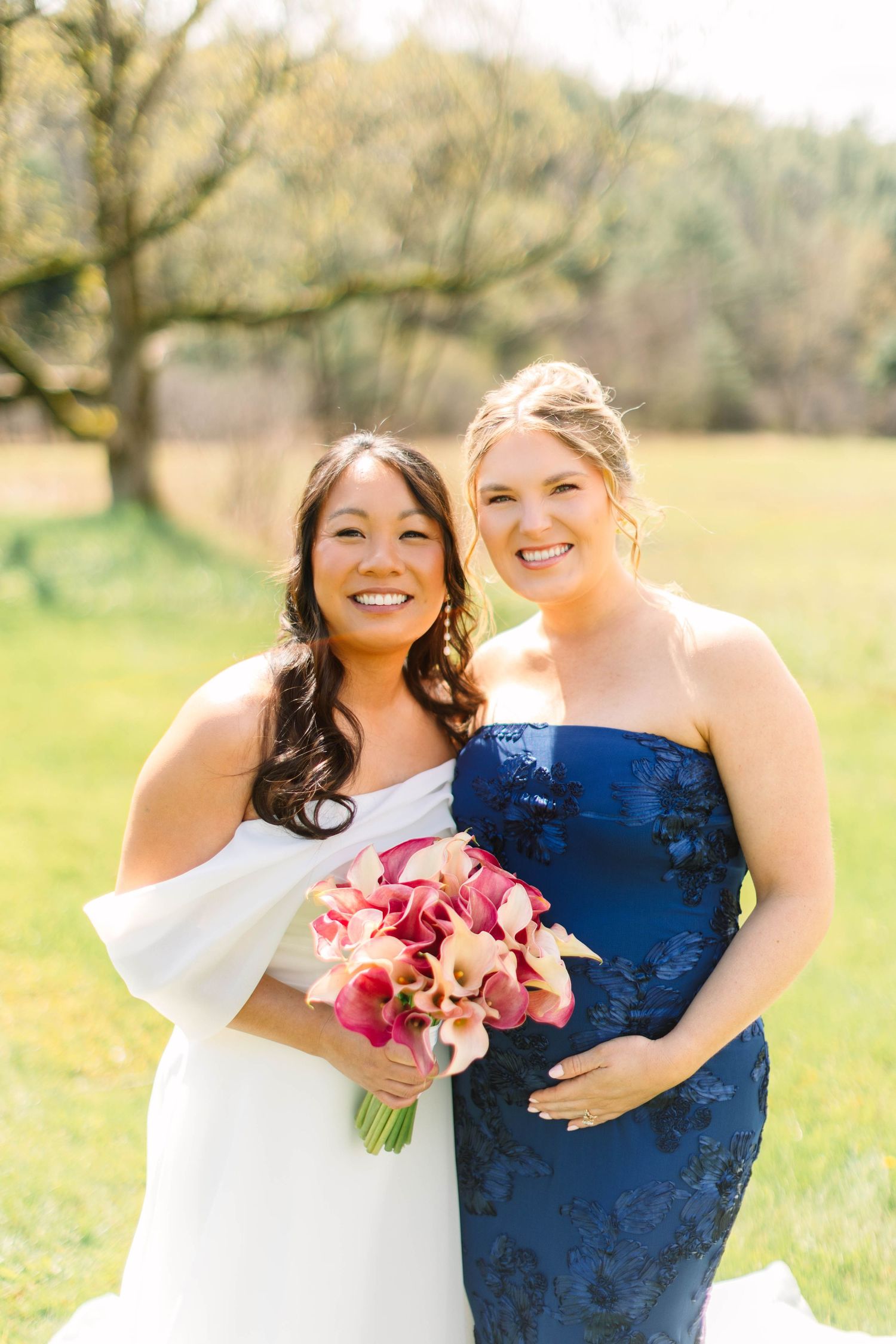 Bride in white off-shoulder gown poses with bridesmaid in navy blue dress holding pink bouquet.