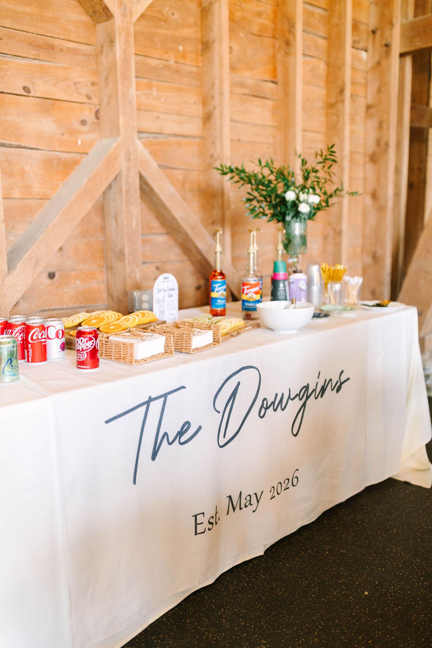 Rustic barn reception table with white tablecloth reading The Dowgins Est. May 2026 with food and drinks.