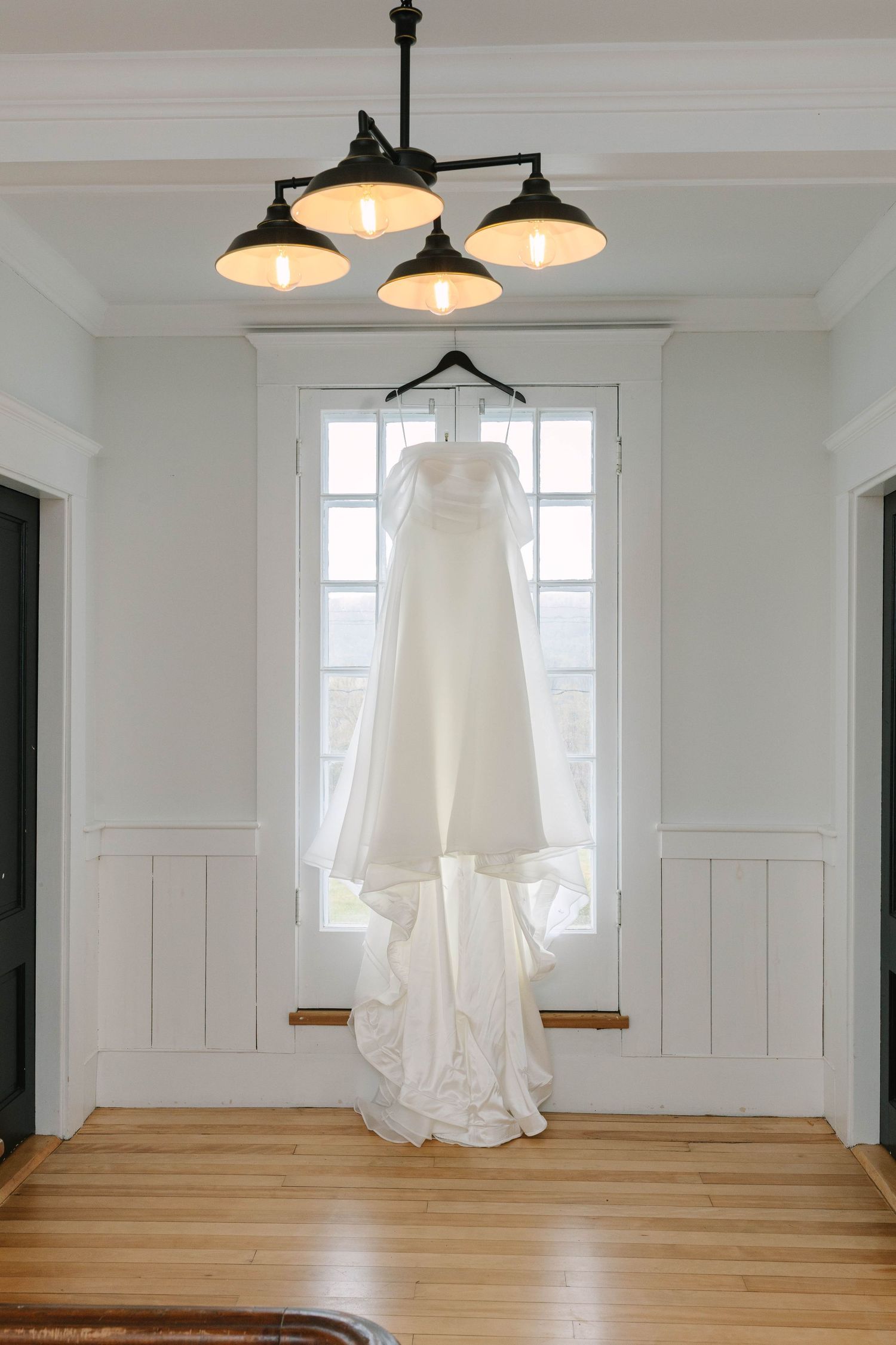 White wedding gown hanging from chandelier in front of bright window in elegant white room with hardwood floors.