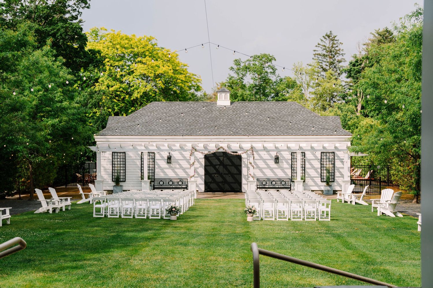 Charming white stone venue with Adirondack chairs on green lawn surrounded by trees.