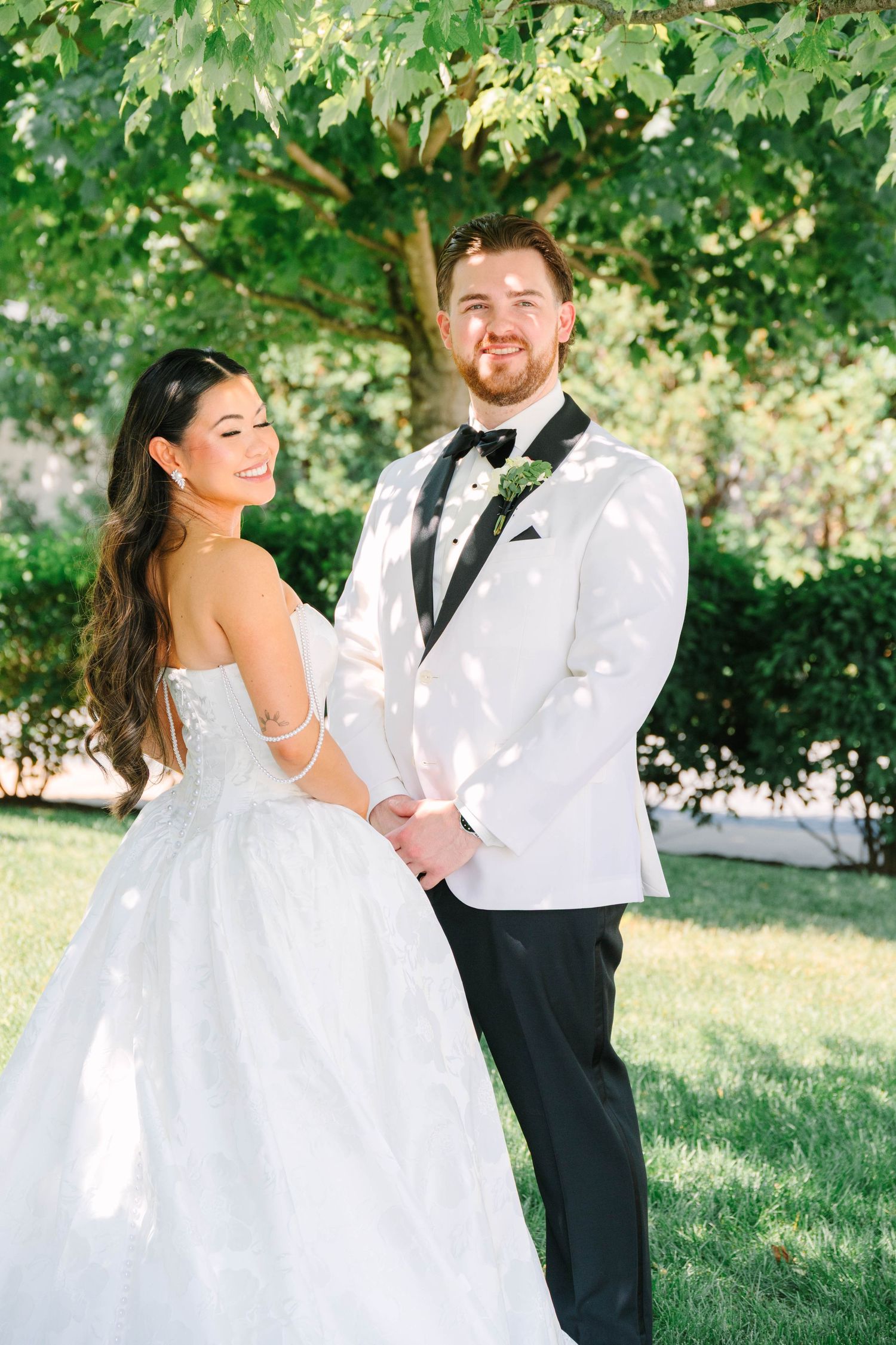 Smiling bride and groom holding hands in sunlit garden, groom wearing white tuxedo with bow tie.