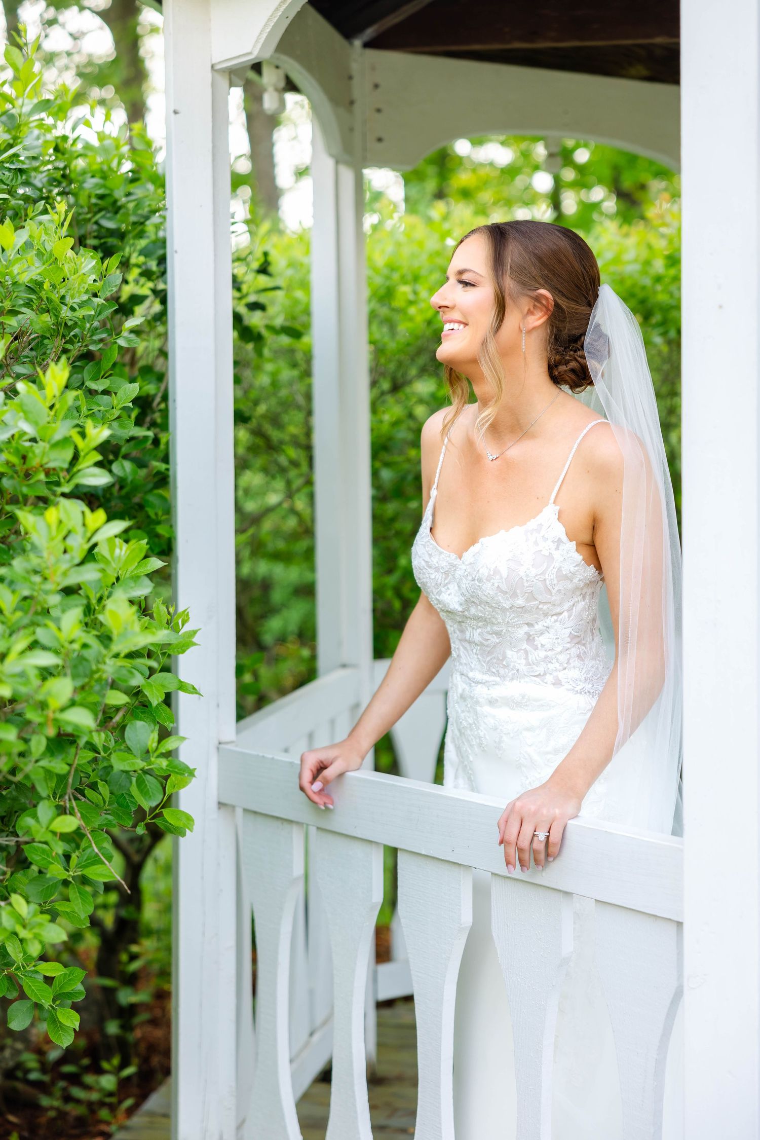Elegant bride in lace wedding dress and veil smiling in white gazebo surrounded by greenery.
