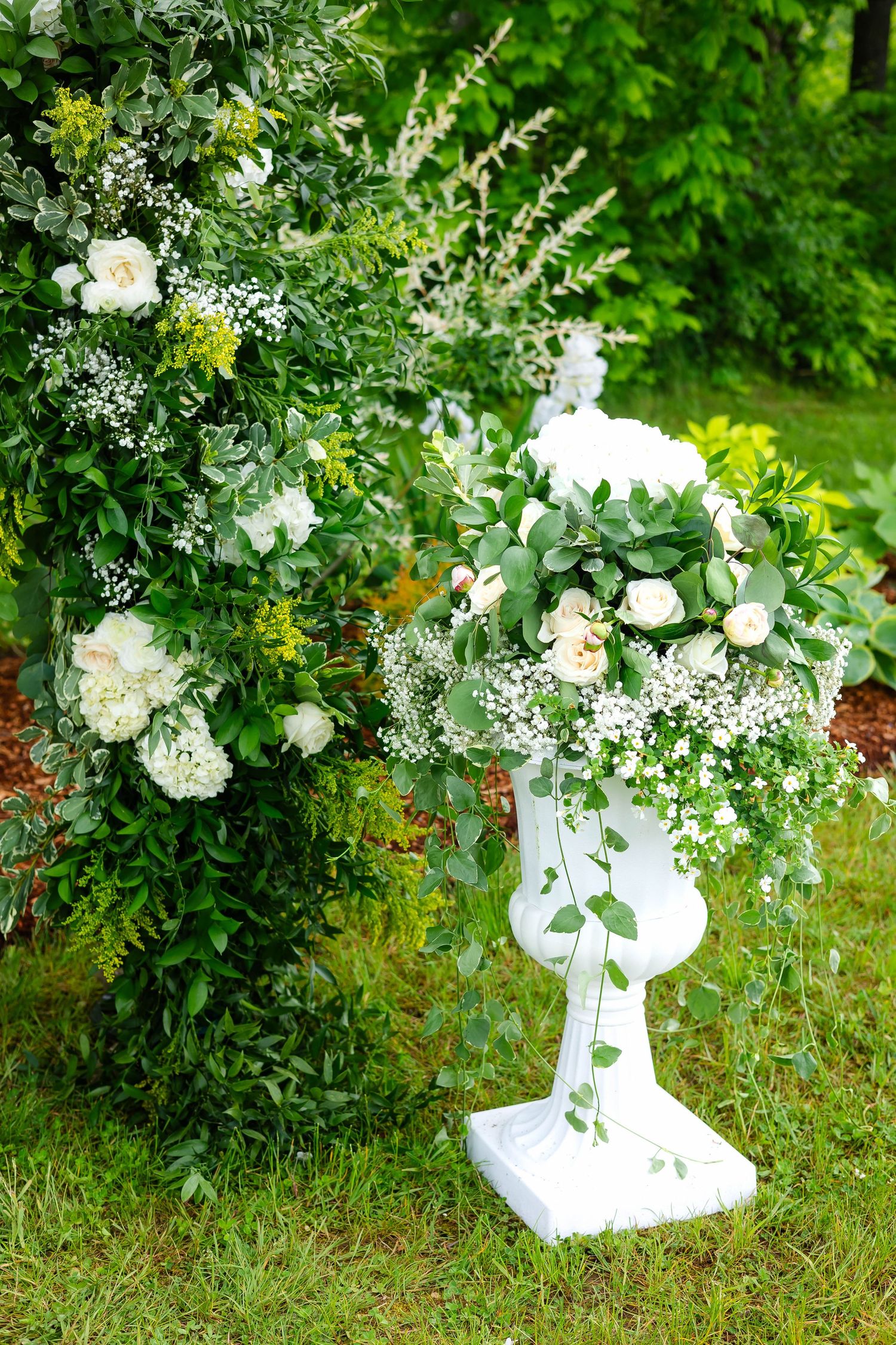 White pedestal urn overflowing with white roses, hydrangeas, and trailing greenery at an outdoor wedding.