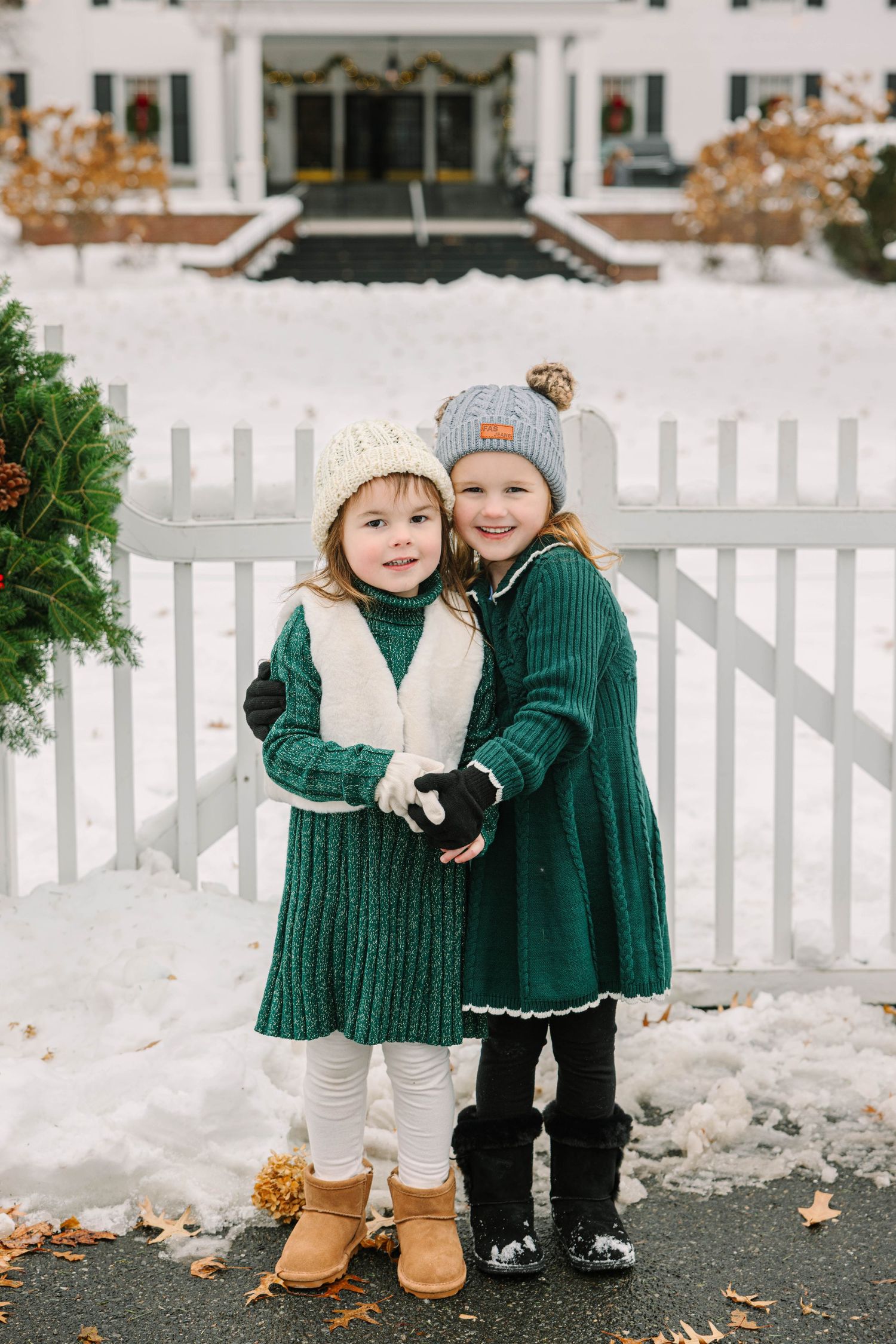 Two little girls in green knit dresses and winter hats hugging outside a snowy white fence.