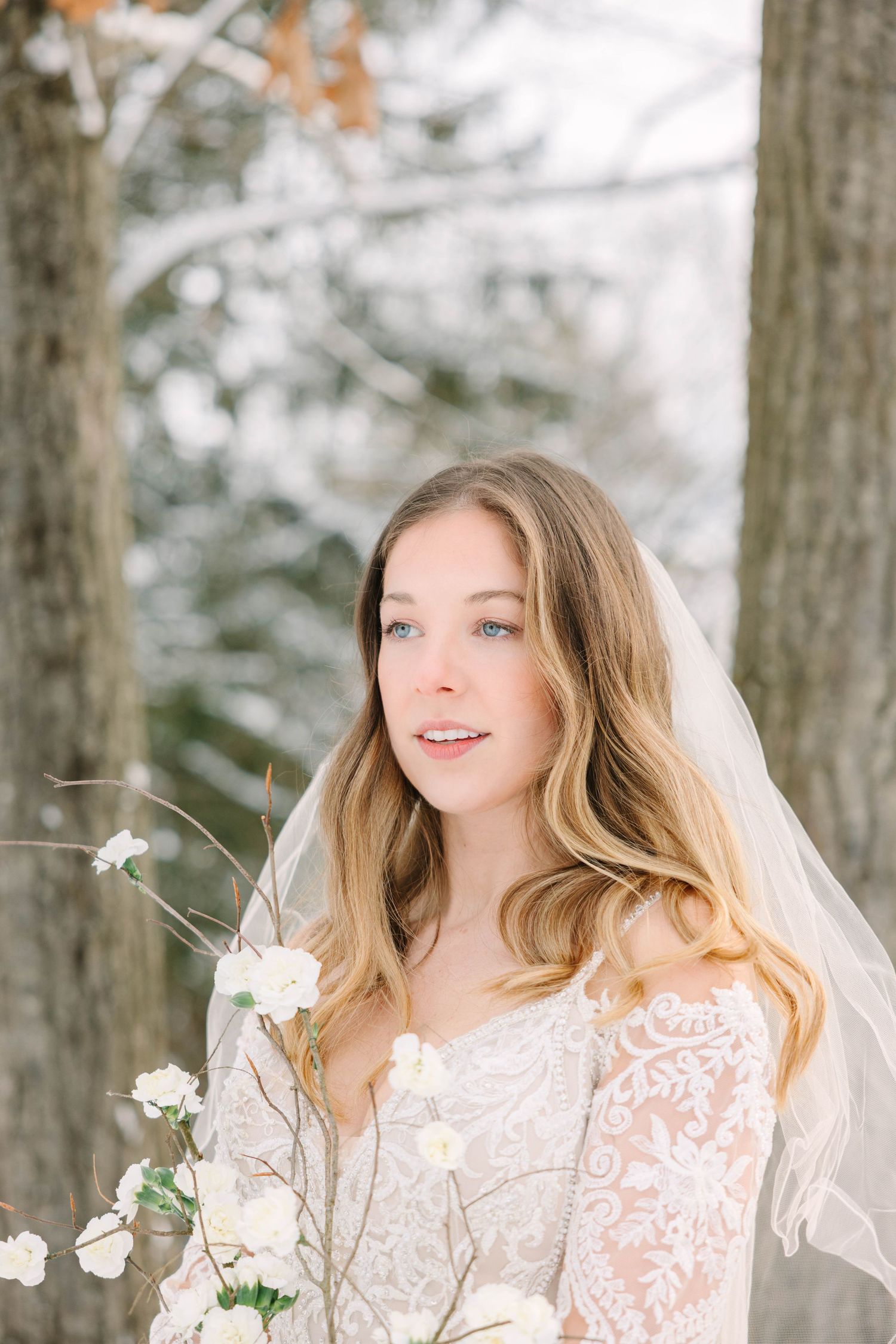 Beautiful blue-eyed bride in lace gown with veil posing among snow-dusted winter trees.