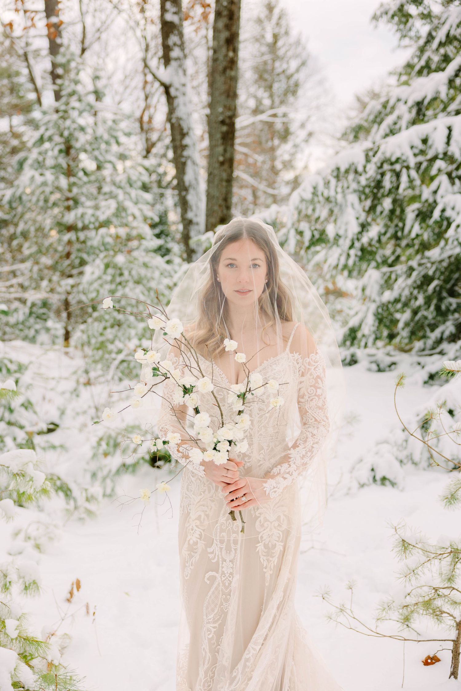Bride in lace dress with veil holding white floral branches in snowy forest.