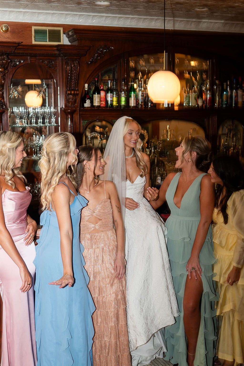 Bride in white wedding gown surrounded by bridesmaids in colorful dresses laughing together at a bar.