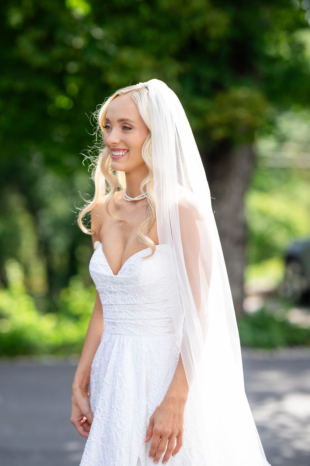 Blonde bride in strapless lace gown and veil smiling outdoors on wedding day.
