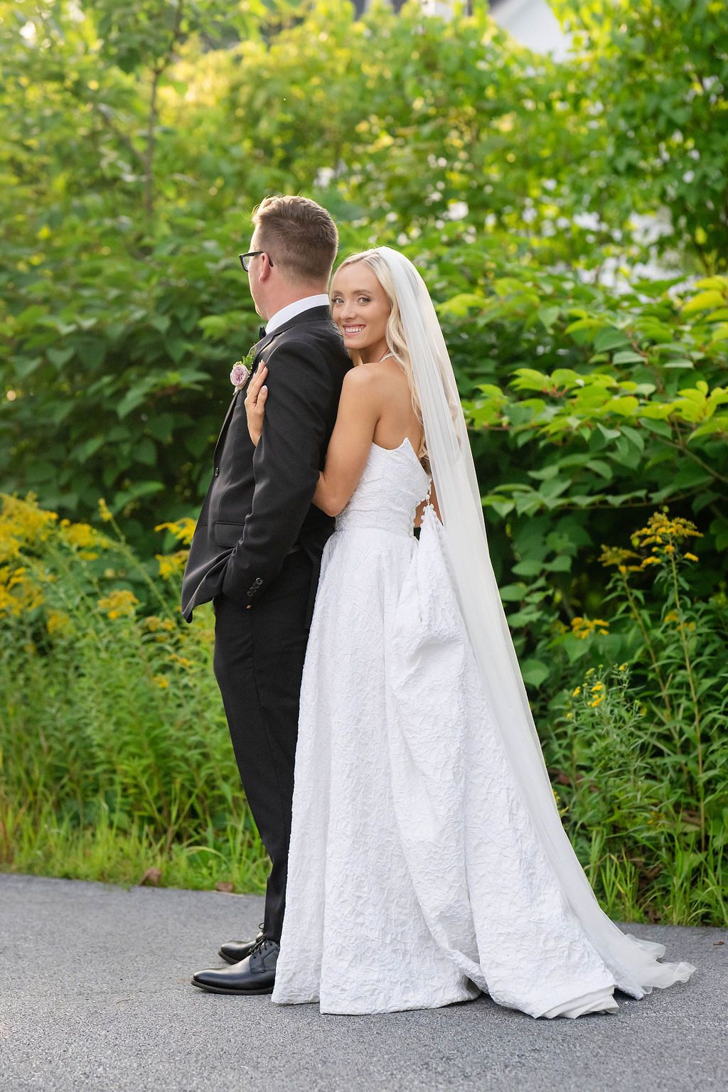 Bride in white lace gown with veil embraces groom in black tuxedo outdoors surrounded by lush greenery.