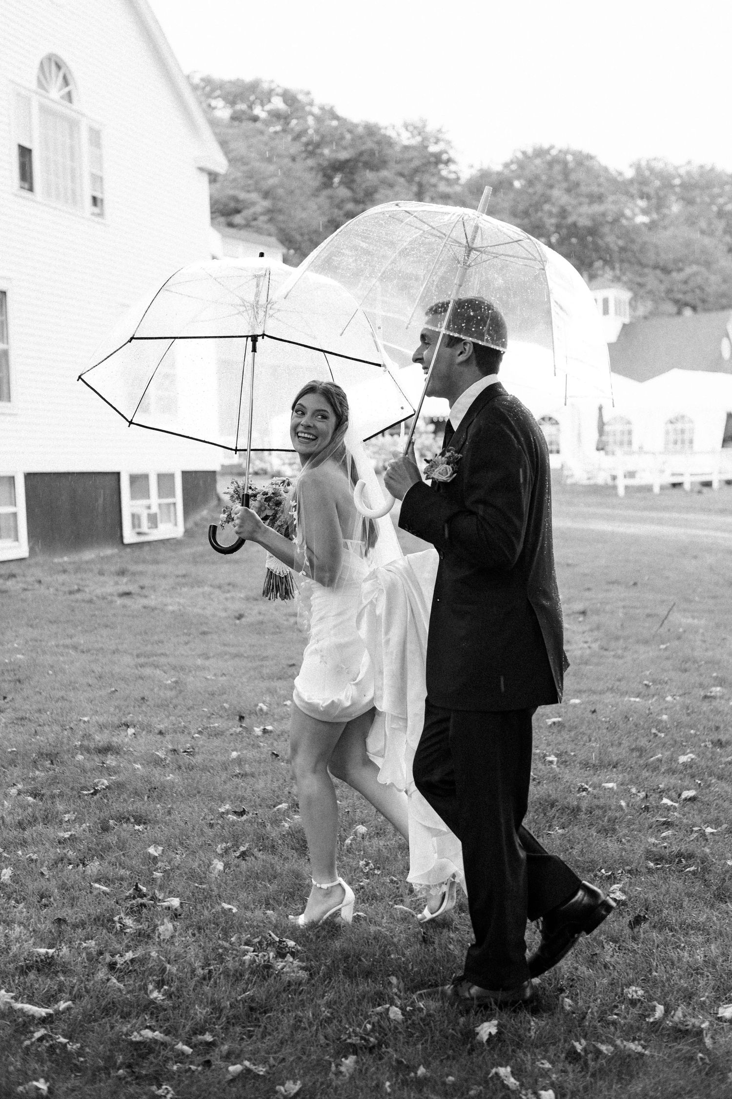 Black and white photo of a smiling bride and groom walking with clear umbrellas in the rain on their wedding day.