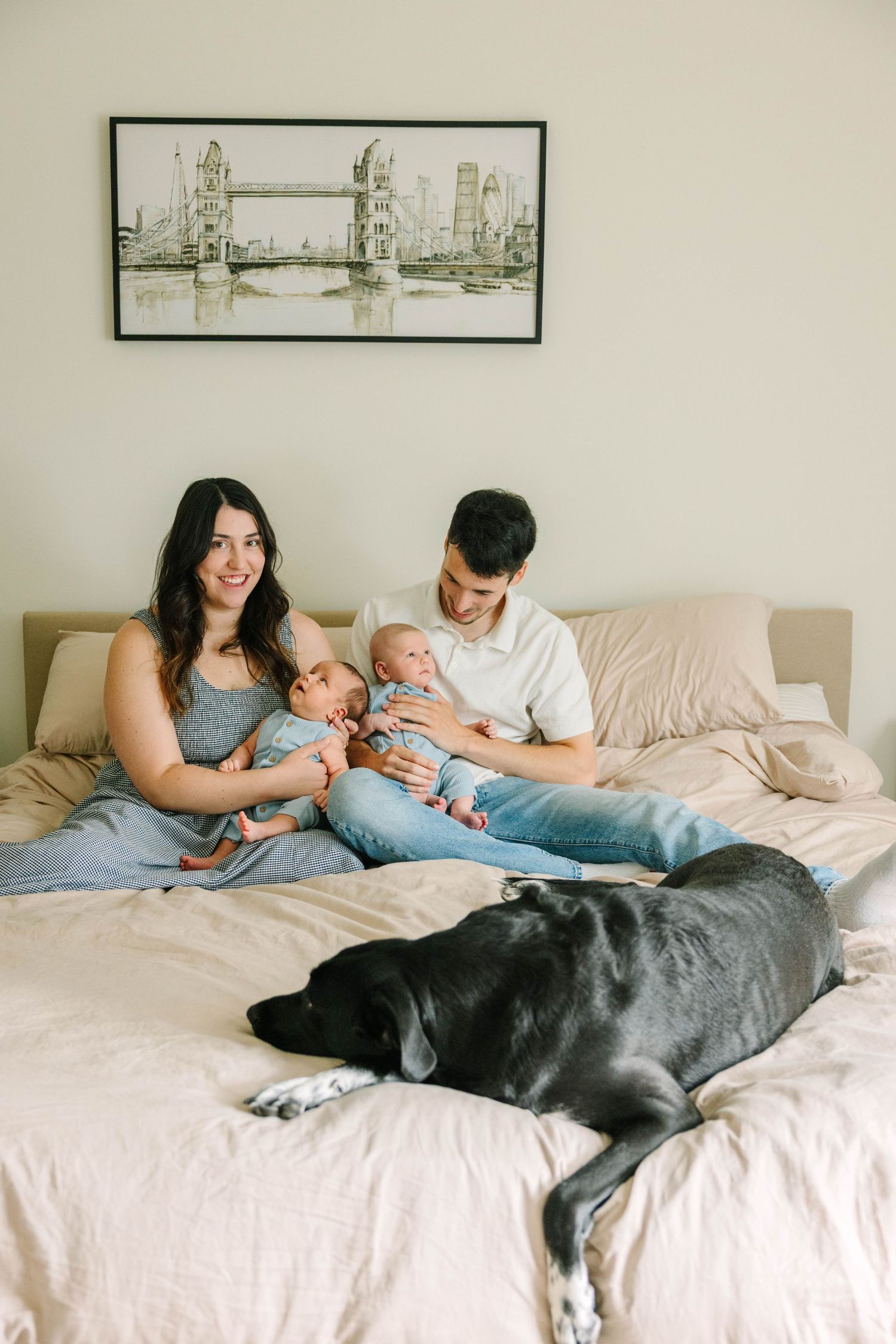 Young family with newborn twins and a black Labrador dog relaxing together on a bed at home.