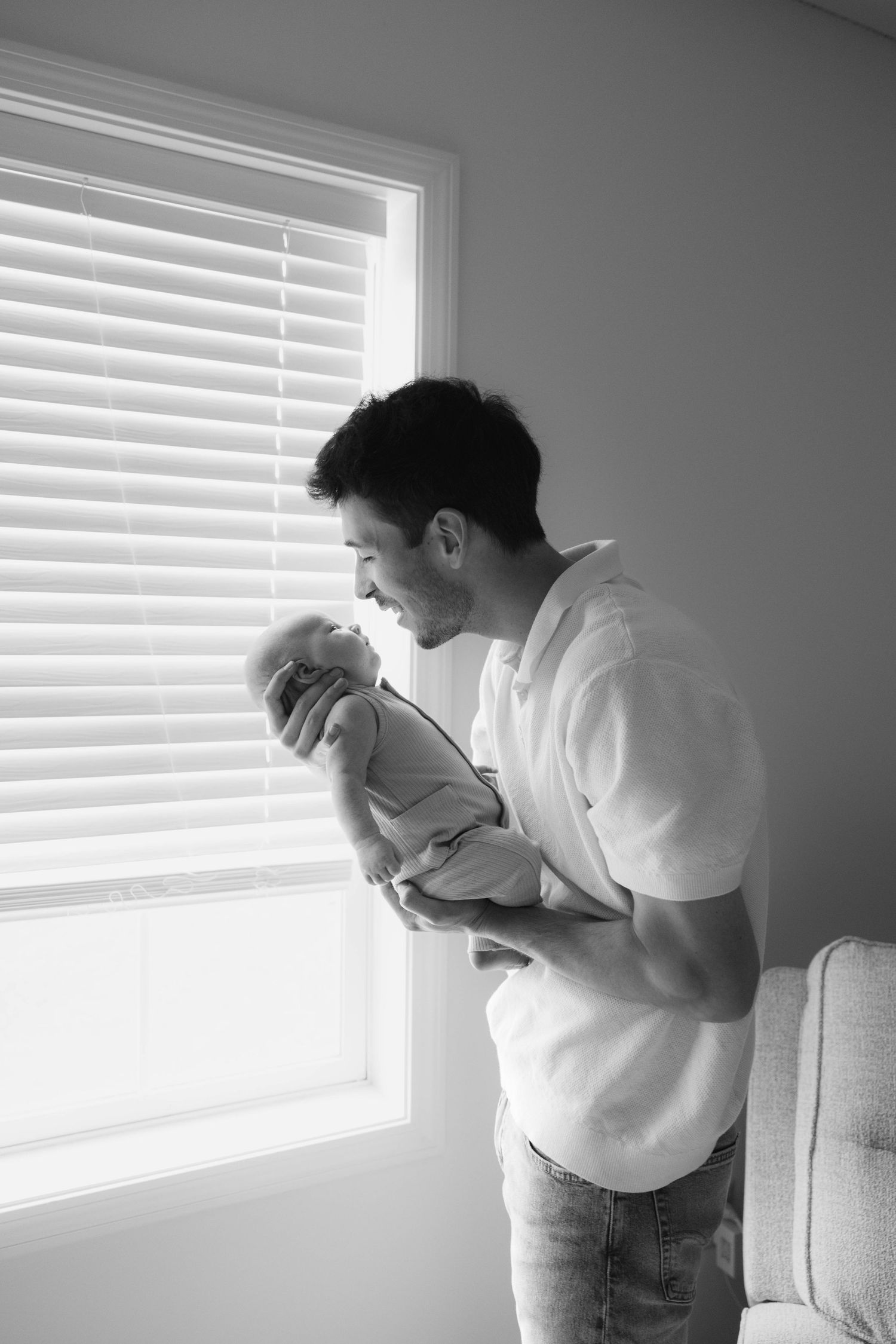 Black and white photo of a father tenderly holding his newborn baby near a bright window with blinds.