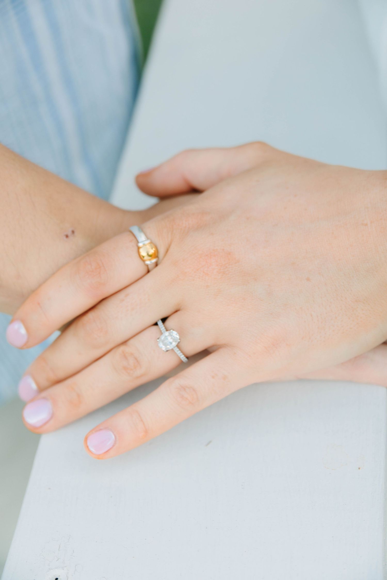 Close-up of two hands wearing elegant rings, one gold and one diamond halo, with soft pink manicured nails.
