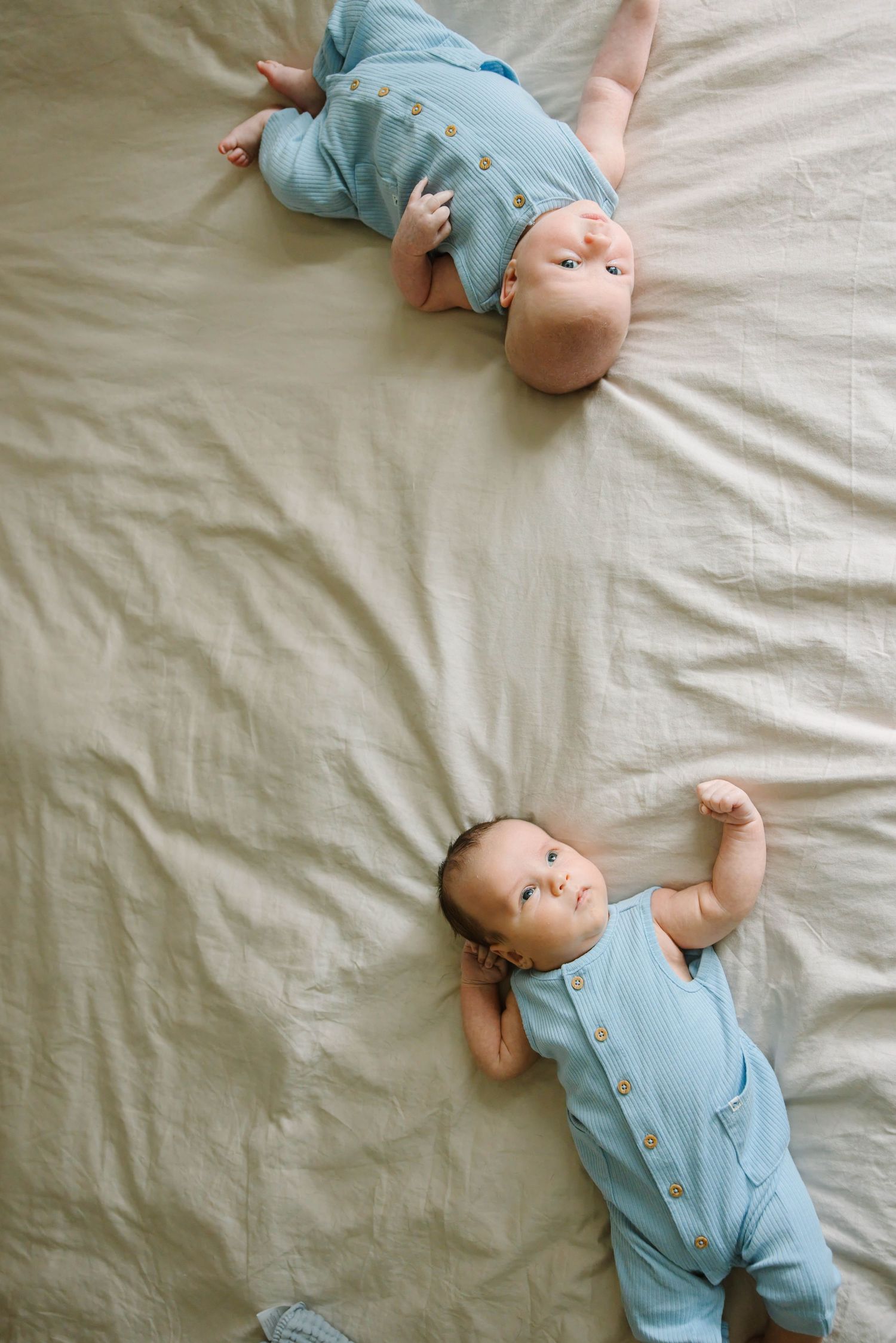 Two babies in blue rompers lying on a beige bed, looking up at the camera.
