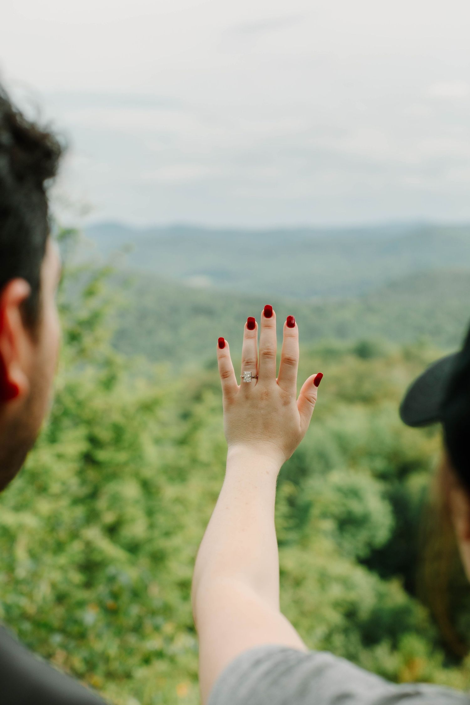 Woman displaying engagement ring with red nails against scenic mountain forest backdrop.